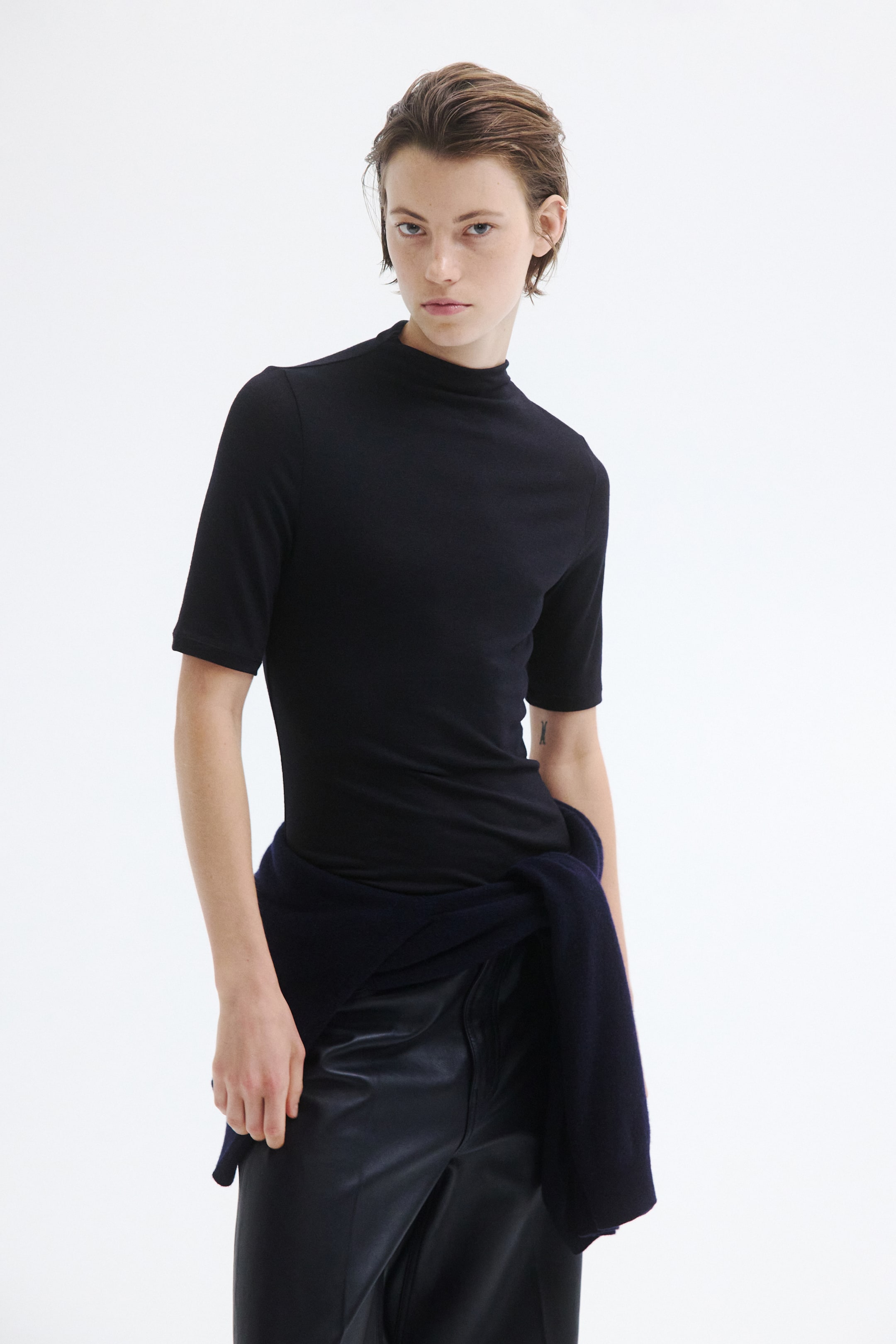 View larger image: The Warming Slim-Fit Mock Turtleneck Top is shown with a slim-fit silhouette from the front, featuring a mock neck and elbow-length sleeves. The black top is styled tucked into dark leather trousers, with a knit jumper tied around the waist.