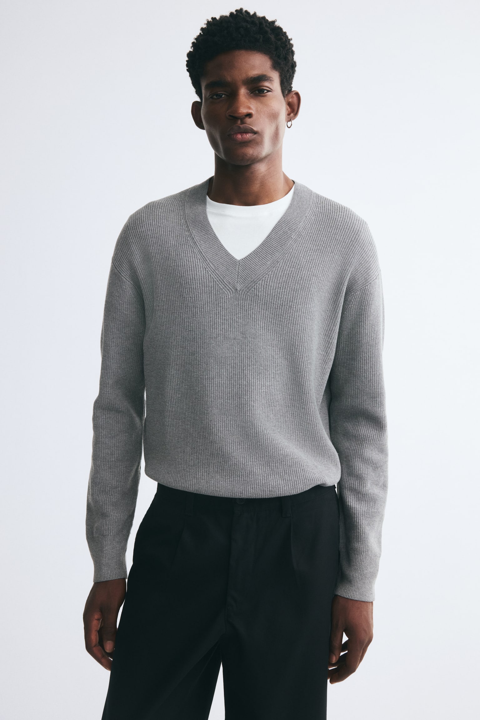 A grey rib-knit sweater featuring a V-neck, dropped shoulders, and long sleeves is worn by the model. The relaxed-fit sweater is layered over a white crew-neck T-shirt and paired with black trousers. - 1