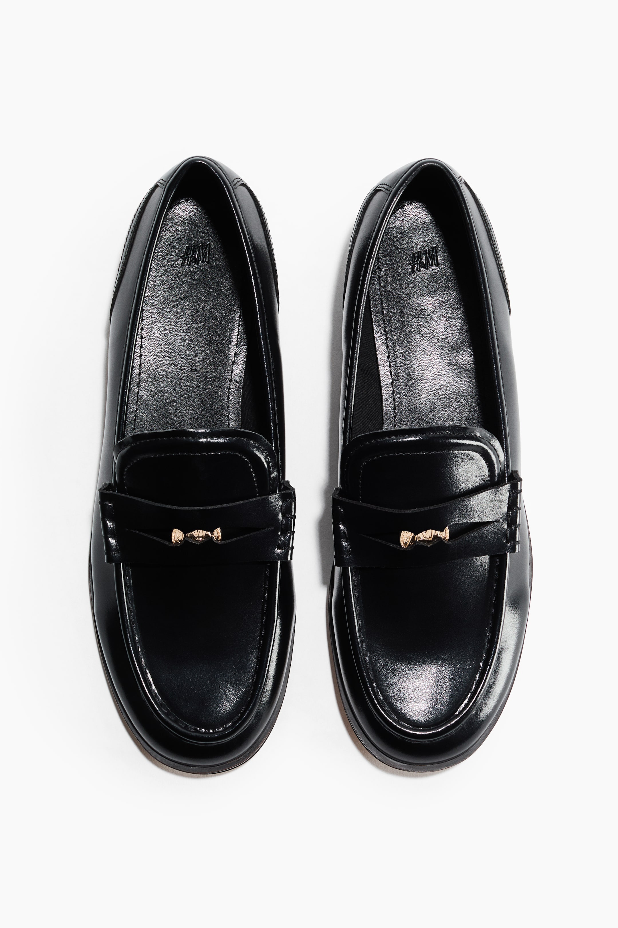 Größeres Bild ansehen: Ein Paar schwarze Loafer steht nach vorne gerichtet, mit einer glatten, glänzenden Oberfläche und Mokassin-Nähten. Jeder Loafer zeigt einen Riemen über dem Spann mit einem goldfarbenen Horsebit-Detail, und das "H&M"-Logo ist auf der Innensohle sichtbar.