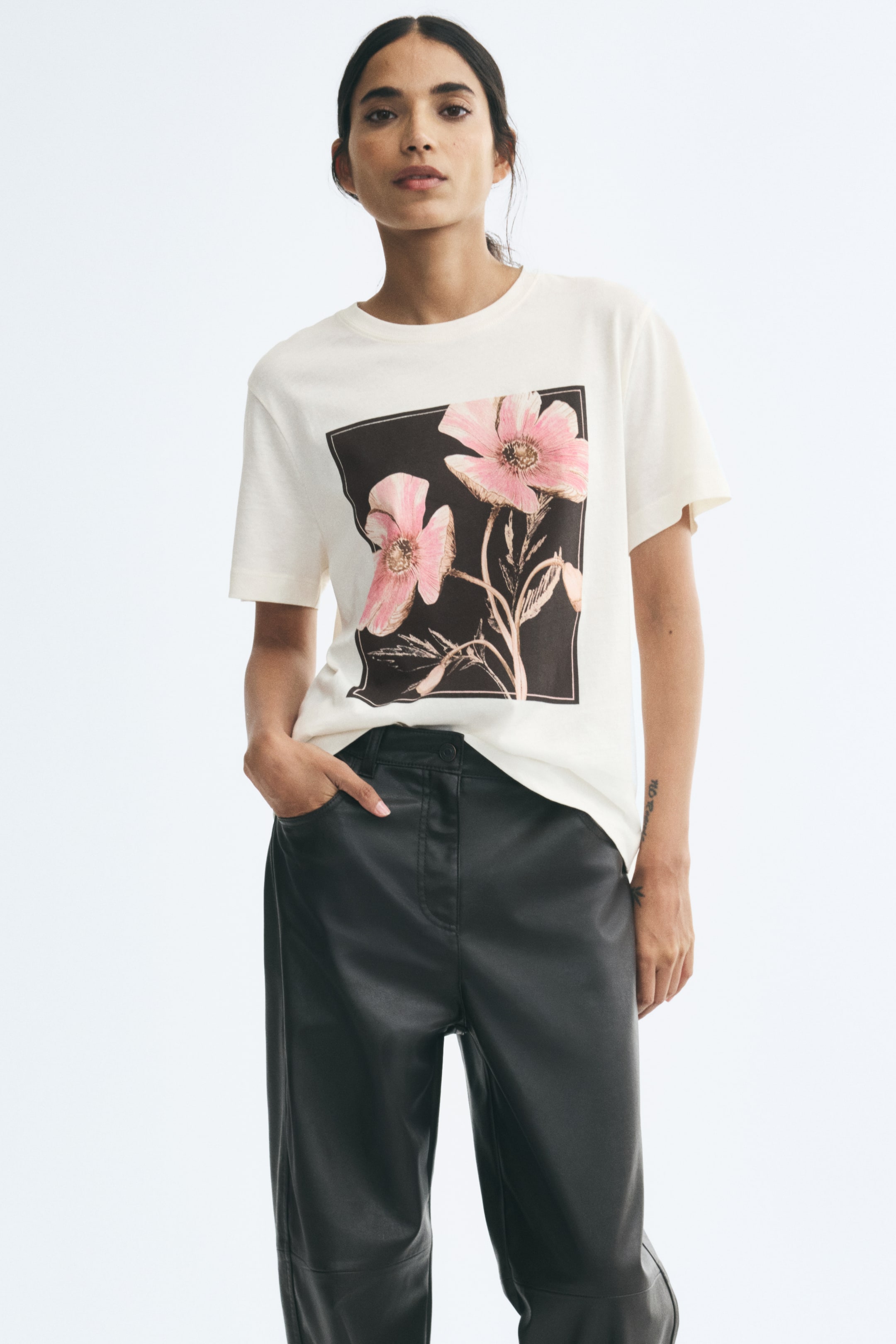 View larger image: An off-white cotton jersey T-shirt featuring a round neckline, dropped shoulders, and a square graphic print of pink flowers on the front is being worn by the model. The T-shirt is styled partially tucked into high-waisted black leather-look trousers.