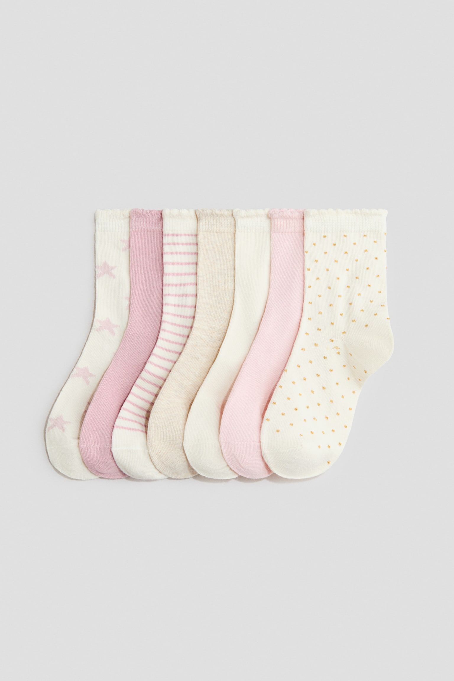 Seven fine-knit socks in a soft cotton blend are laid flat in an overlapping row, showing their front profiles with designs including stars, stripes, solid pastel shades, and polka dots. Each sock features a scalloped elasticated top cuff and a shaped heel and toe.