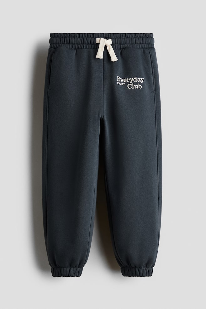 Dark gray sweatshirt joggers are presenting from the FRONT, with an elasticated waist featuring an off-white drawstring tie. White text reading 'Everyday Enjoy Club' is embroidered on the upper left side, and covered elastication is shaping the hems. - 1
