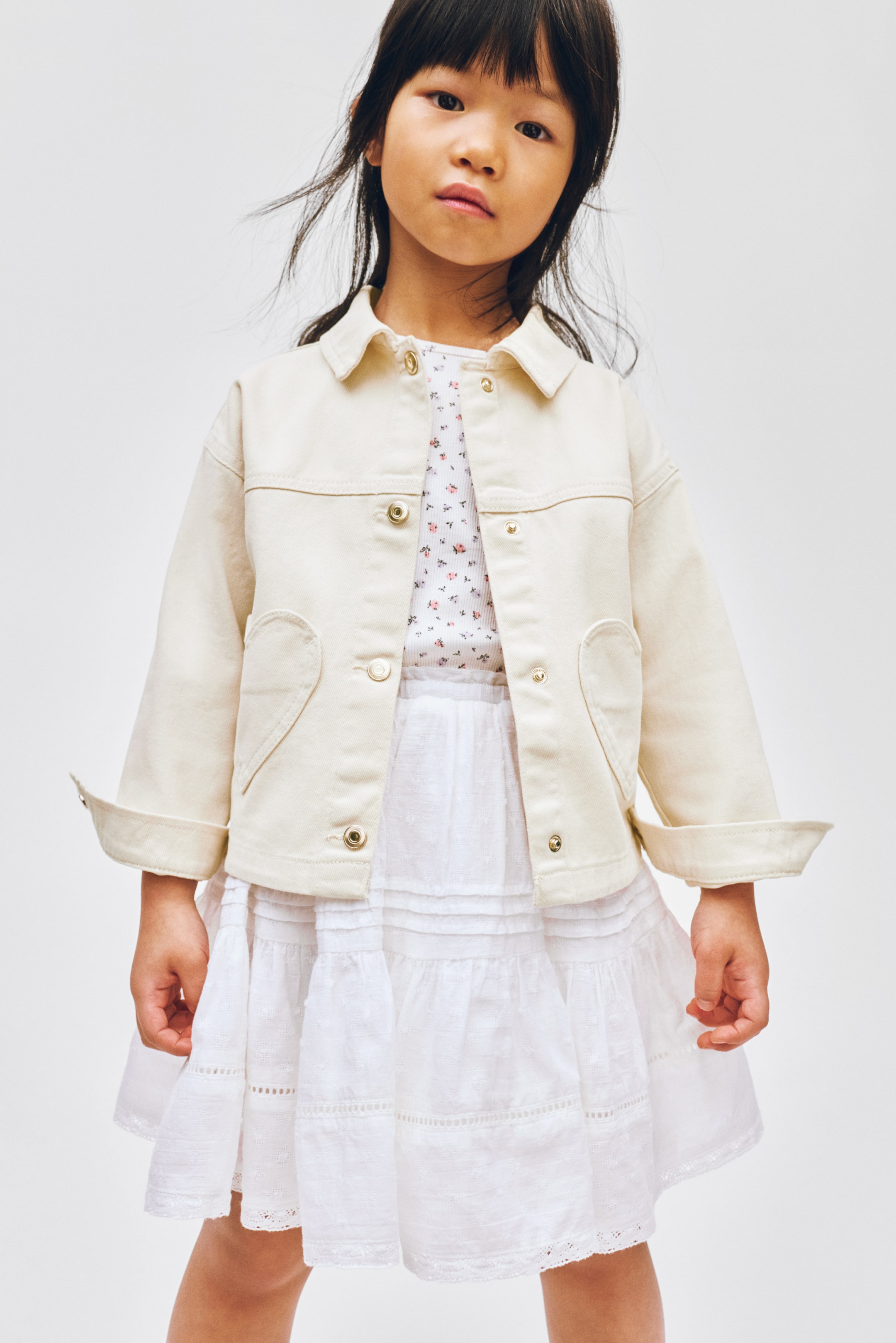 Ver imagen más grande: Una chaqueta de color crema está orientada hacia adelante, con un cuello en punta, cierres de botones de presión dorados y dos bolsillos de parche en forma de corazón en la parte inferior delantera. Está diseñada con costuras visibles y mangas largas que están dobladas en los puños.