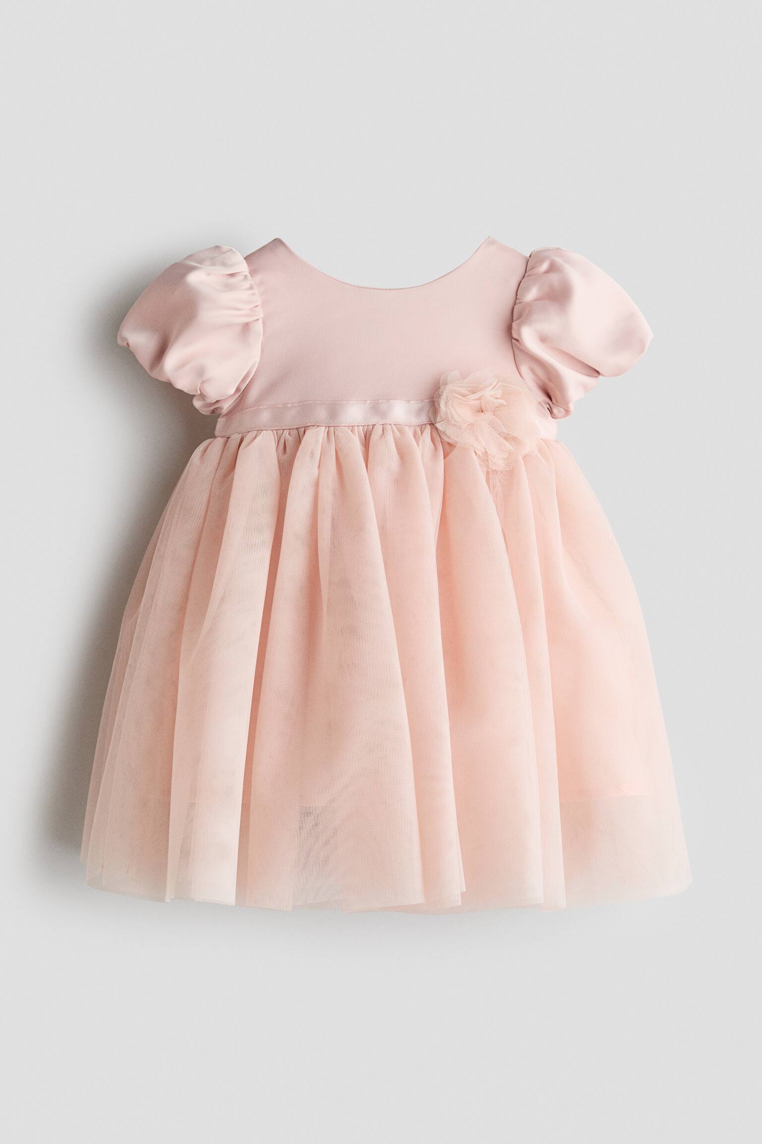 A light pink baby dress is facing front, displaying a smooth satin bodice with short puffed sleeves and a round neckline. A gathered, layered tulle skirt extends from a satin waistband featuring a small fabric flower appliquÃ©.