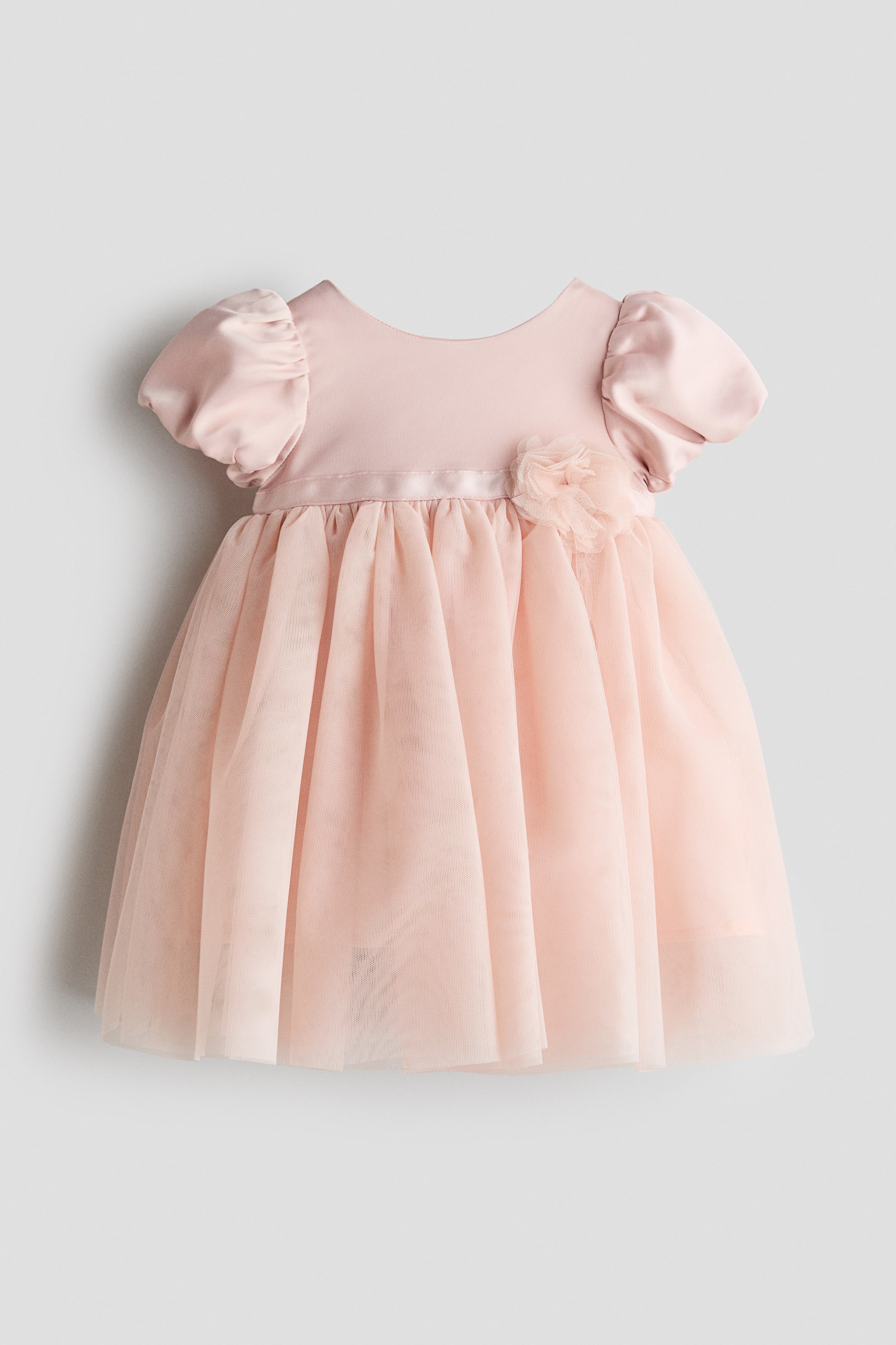 View larger image: A light pink baby dress is facing front, displaying a smooth satin bodice with short puffed sleeves and a round neckline. A gathered, layered tulle skirt extends from a satin waistband featuring a small fabric flower appliqué.
