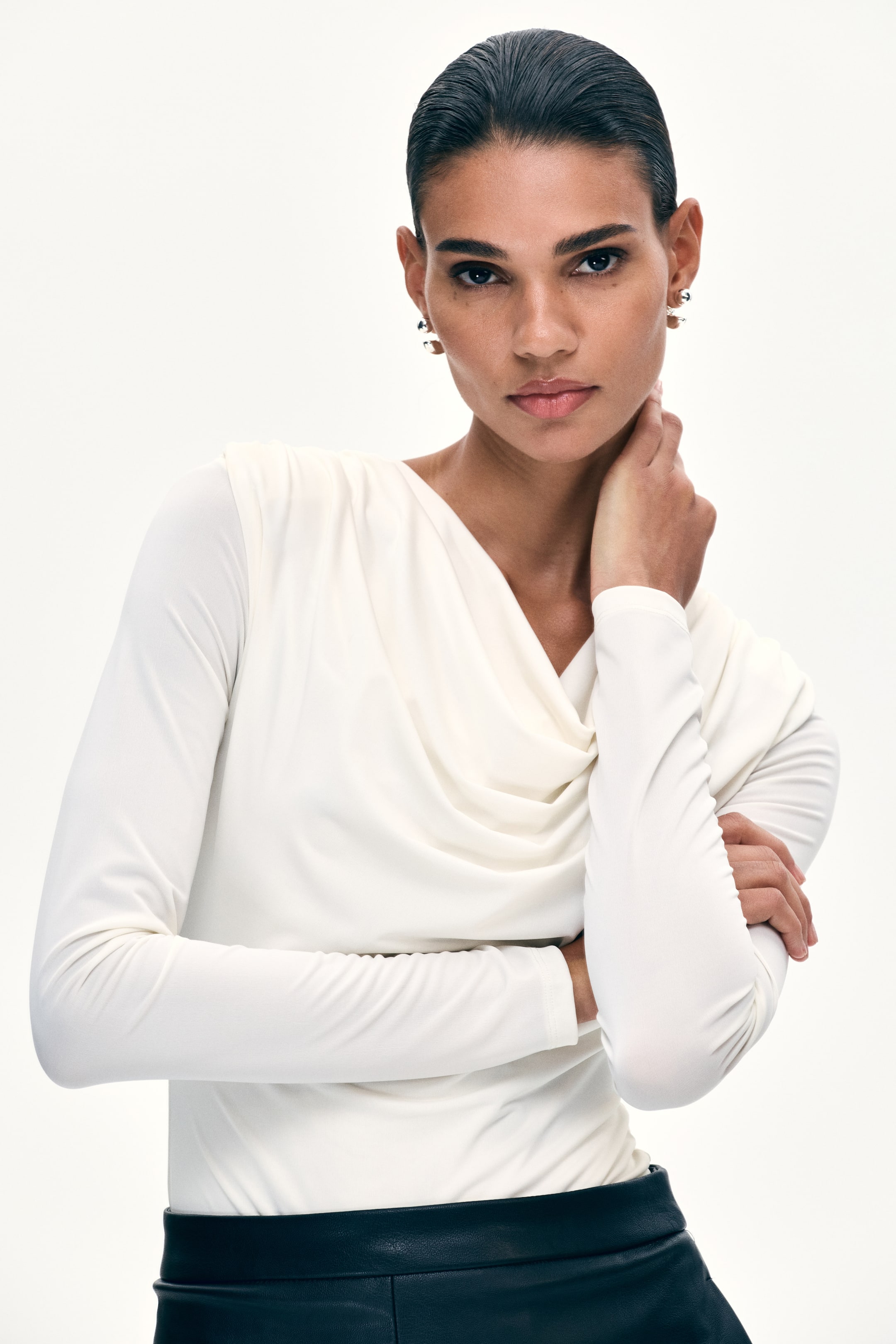 View larger image: The model is wearing a cream long-sleeved top featuring a draped cowl neck. She is seen touching her neck and crossing her arms.
