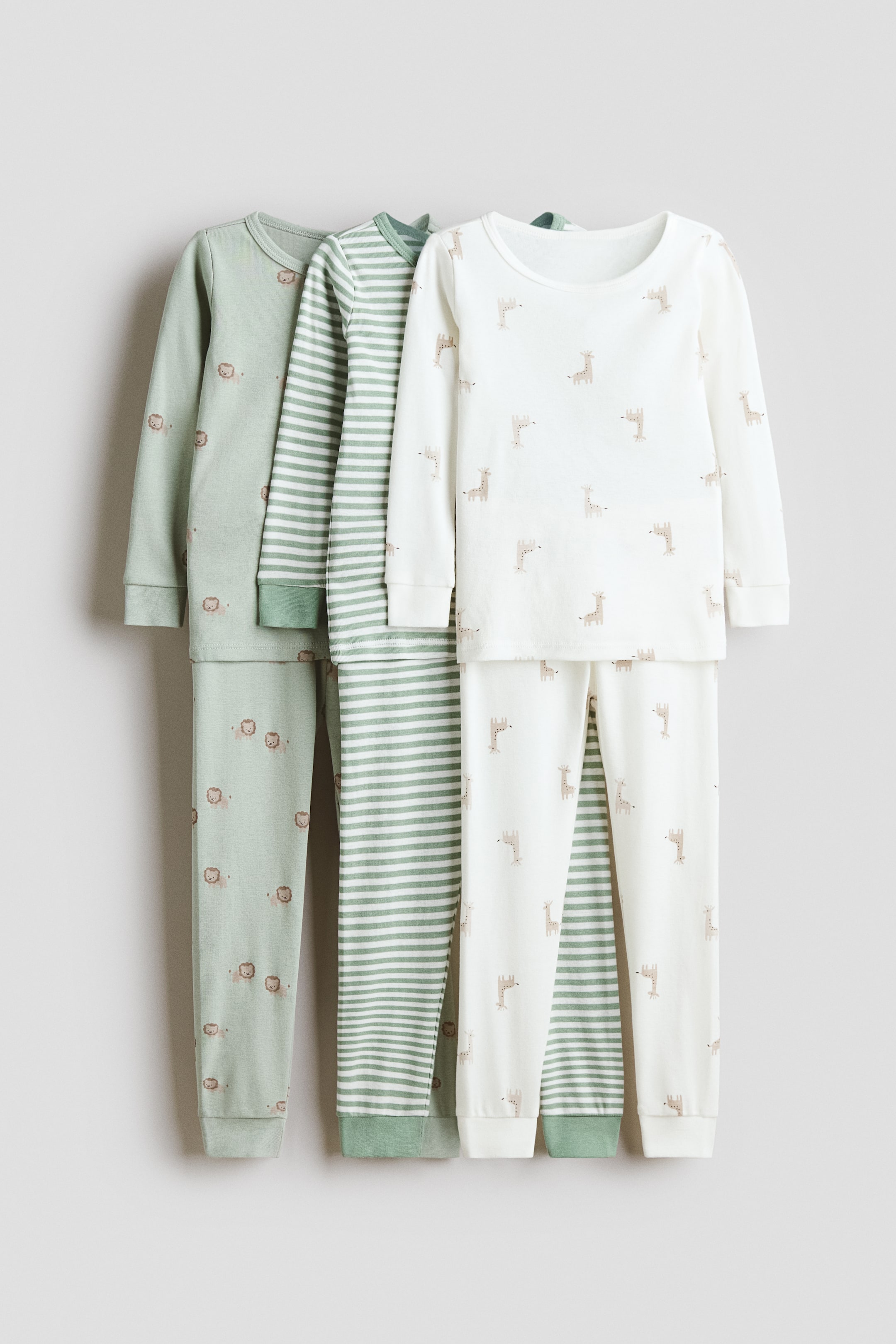 View larger image: Three children's pajama sets are displayed with their long-sleeved tops and matching long pants featuring ribbed cuffs. The leftmost set is light green with repeating lion motifs, the middle set is green and white striped, and the rightmost set is off-white with repeating giraffe motifs.