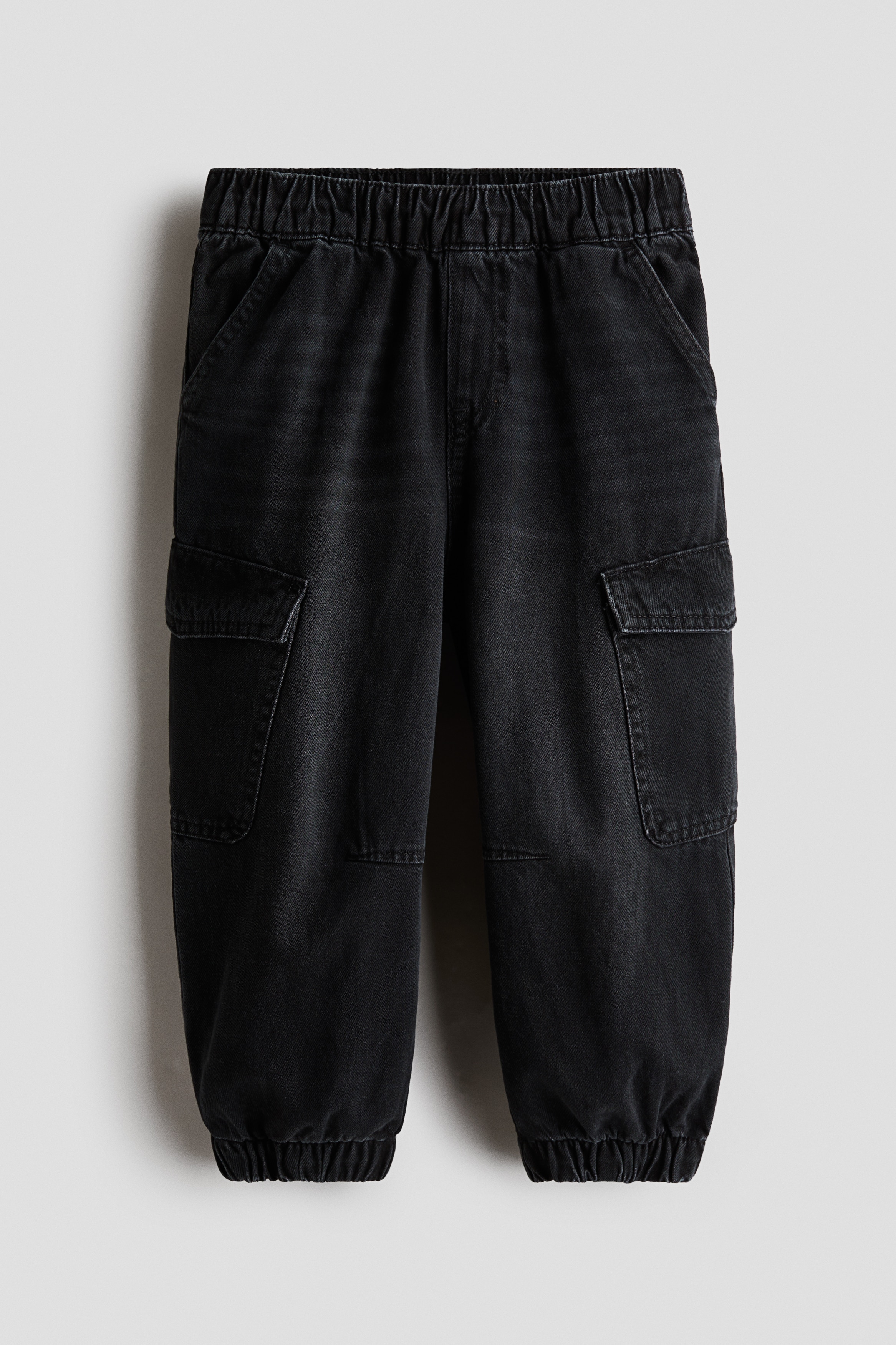 The model is wearing joggers in black washed cotton denim, featuring balloon legs with elasticated hems and side flap pockets. The joggers are styled with a red, white and black colour-blocked zip-up jacket and black trainers.