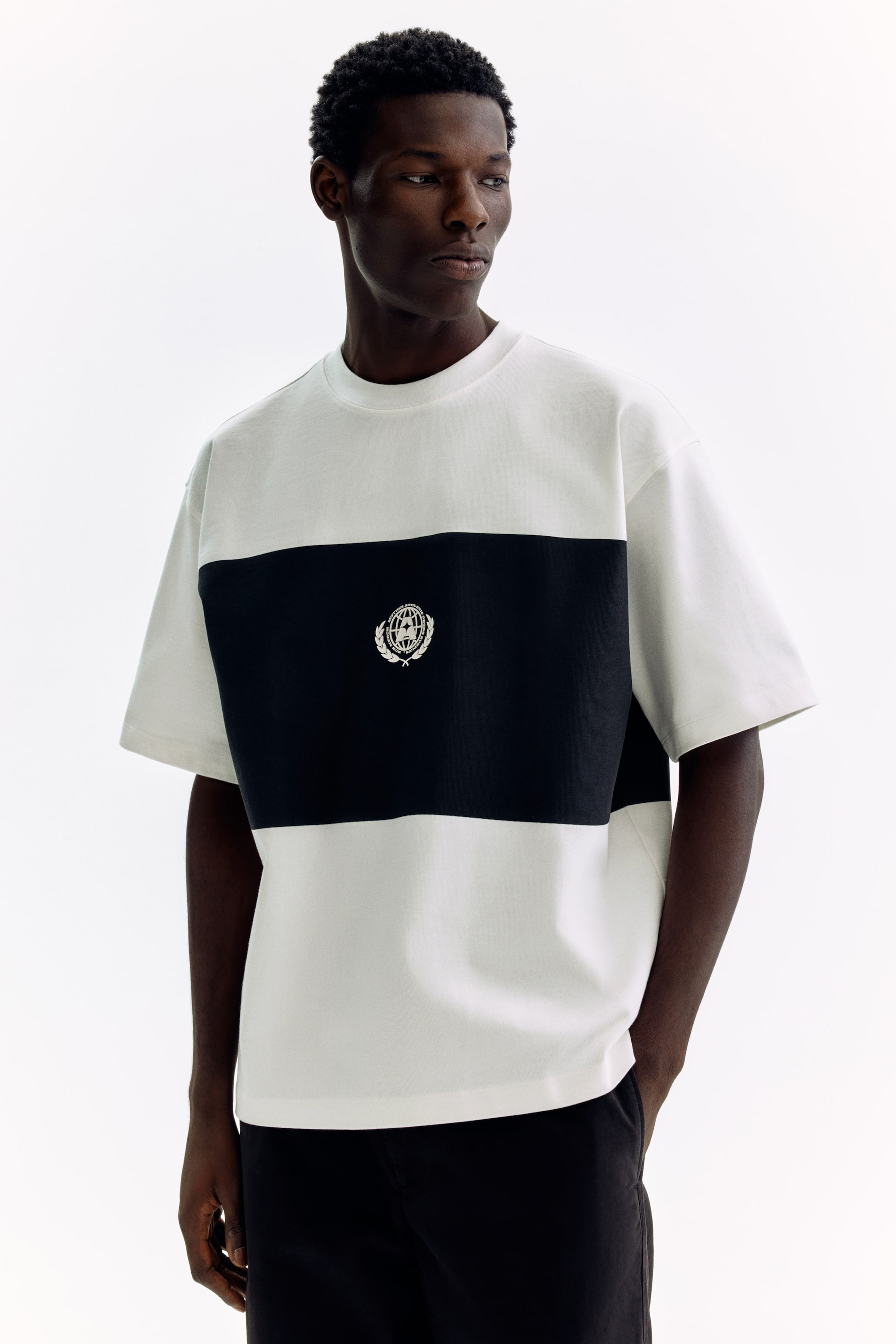 View larger image: This t-shirt showcases a white upper and lower section, divided by a horizontal black stripe across the chest. At the center of the stripe, there's a white embroidered emblem of a globe encircled by laurels. It features a relaxed fit, short sleeves, and a crew neck, perfectly blending style with comfort.
