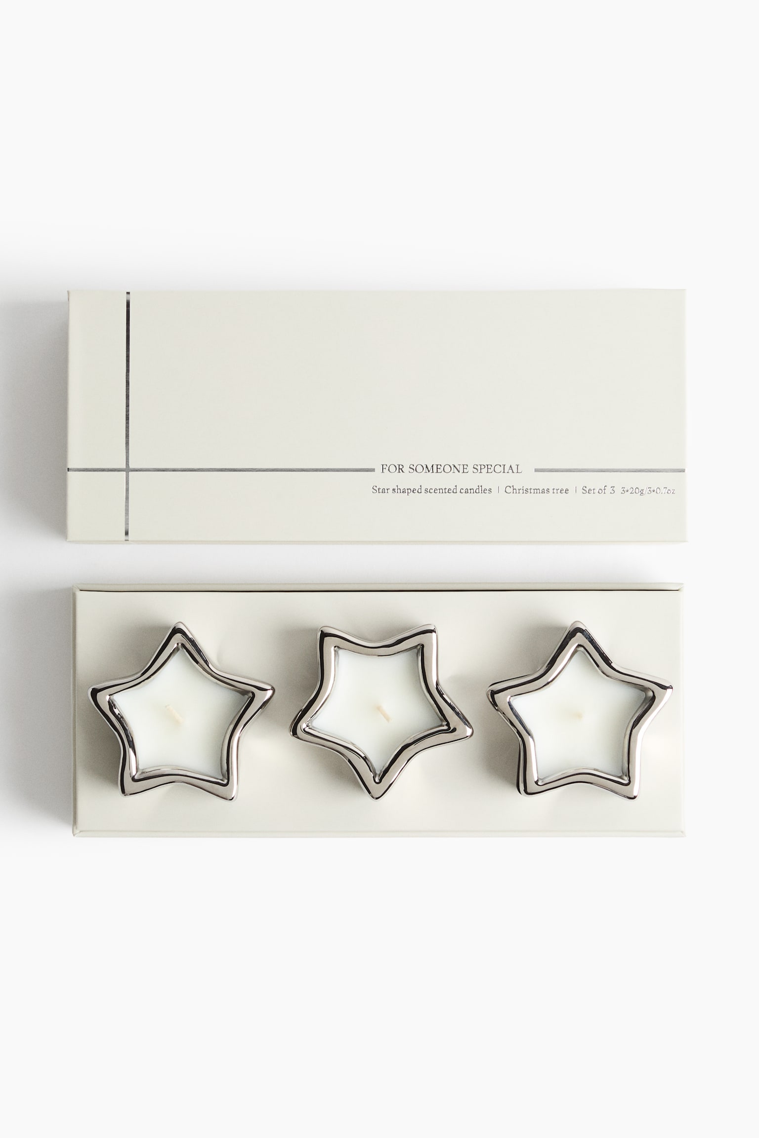 A set of three star-shaped candles featuring white wax and polished silver-toned rims is resting inside an open, cream-colored rectangular box, shown from a front-facing, top-down perspective. The matching cream-colored lid, decorated with thin silver lines and minimalist text, is lying directly above the open box.