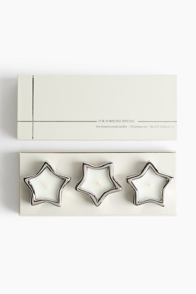 Un ensemble de trois bougies en forme dâÃ©toile, en cire blanche avec des bordures polies ton argent, repose dans une boÃ®te rectangulaire ouverte de couleur crÃ¨me, prÃ©sentÃ© en perspective frontale plongeante. Le couvercle assorti de couleur crÃ¨me, dÃ©corÃ© de fines lignes argentÃ©es et dâun texte minimaliste, est placÃ© juste au-dessus de la boÃ®te ouverte. - 1