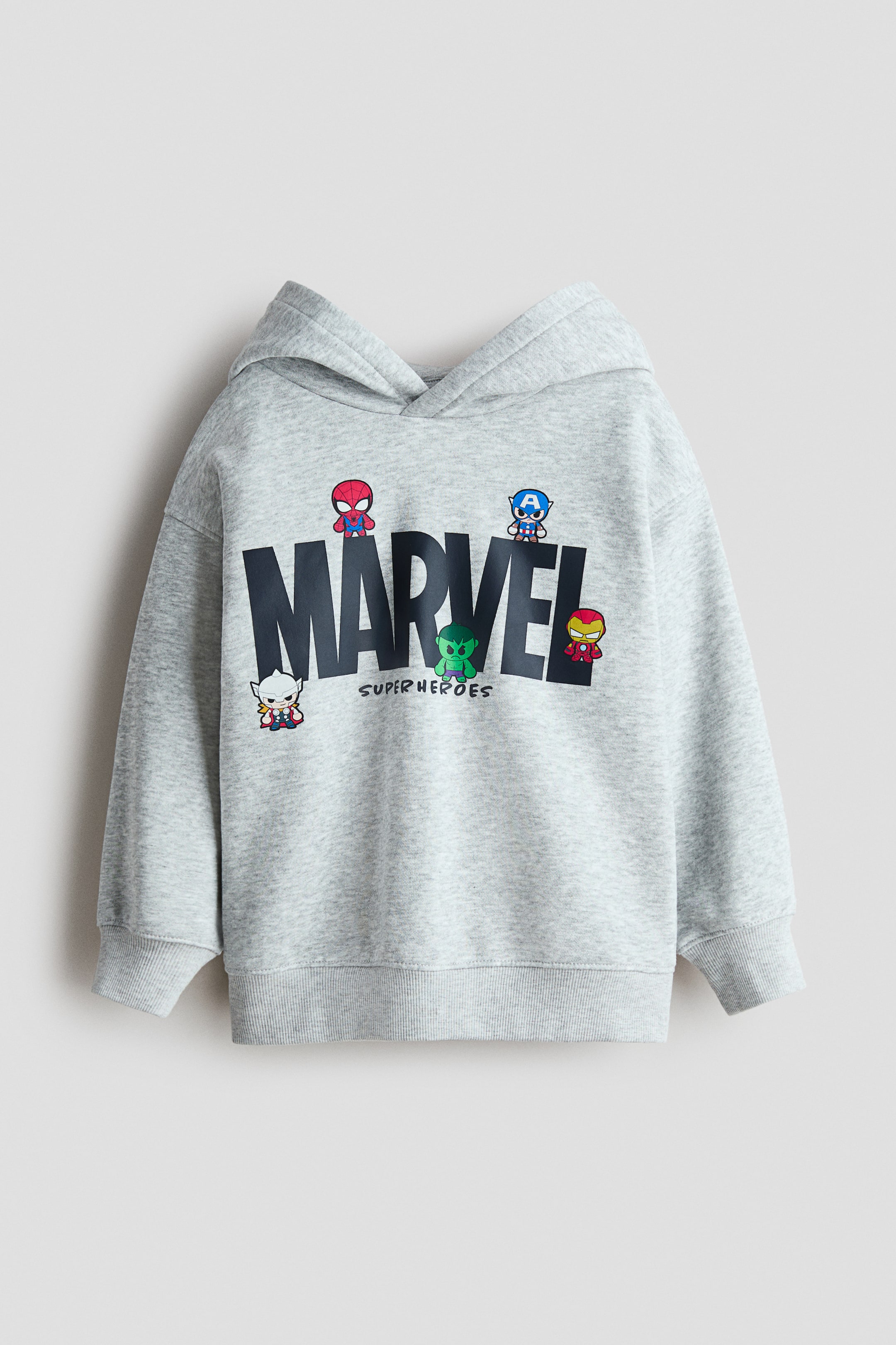 Visa större bild: En löst sittande grå huvtröja i sweatshirt-tyg visar sin framsida, med ett "MARVEL SUPER HEROES"-tryck med tecknade figurmotiv. Den är designad med en fodrad huva som har en omlottdetalj framtill, nedhasade axlar, långa ärmar samt ribbade muddar och fåll.