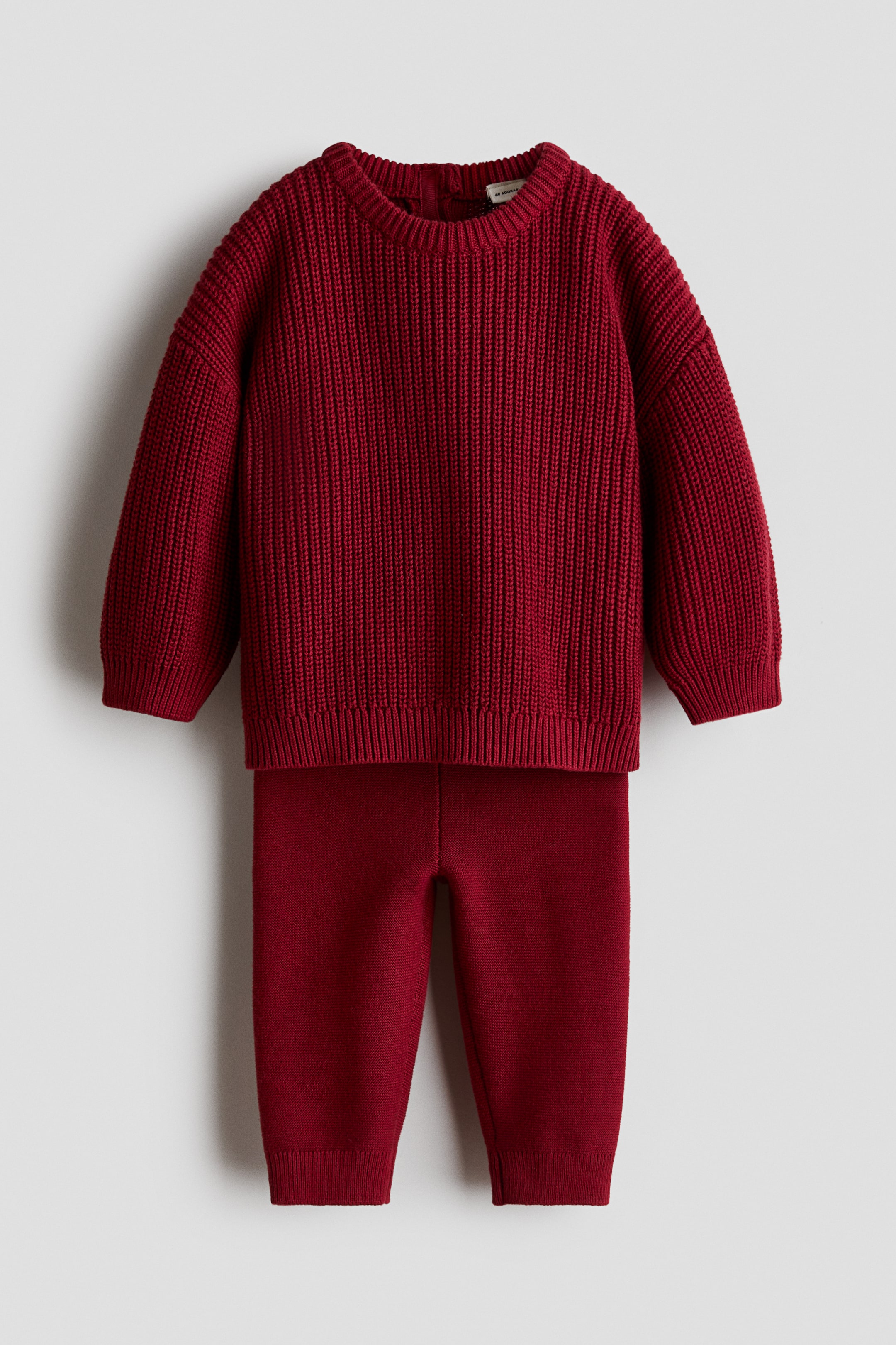 View larger image: A two-piece knit set in deep red is shown from the front, consisting of a patent-knit sweater and matching fine-knit leggings. The sweater features a round neckline, dropped shoulders, and ribbed trim at the collar, cuffs, and hem, while the leggings display a slim fit with ribbed ankle cuffs.