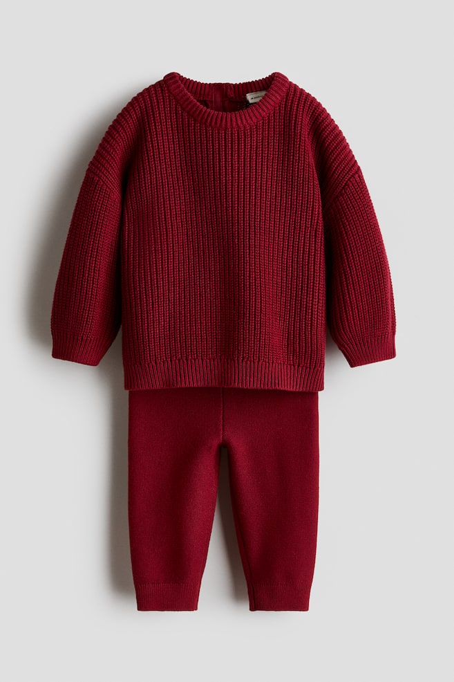 A two-piece knit set in deep red is shown from the front, consisting of a patent-knit sweater and matching fine-knit leggings. The sweater features a round neckline, dropped shoulders, and ribbed trim at the collar, cuffs, and hem, while the leggings display a slim fit with ribbed ankle cuffs. - 1