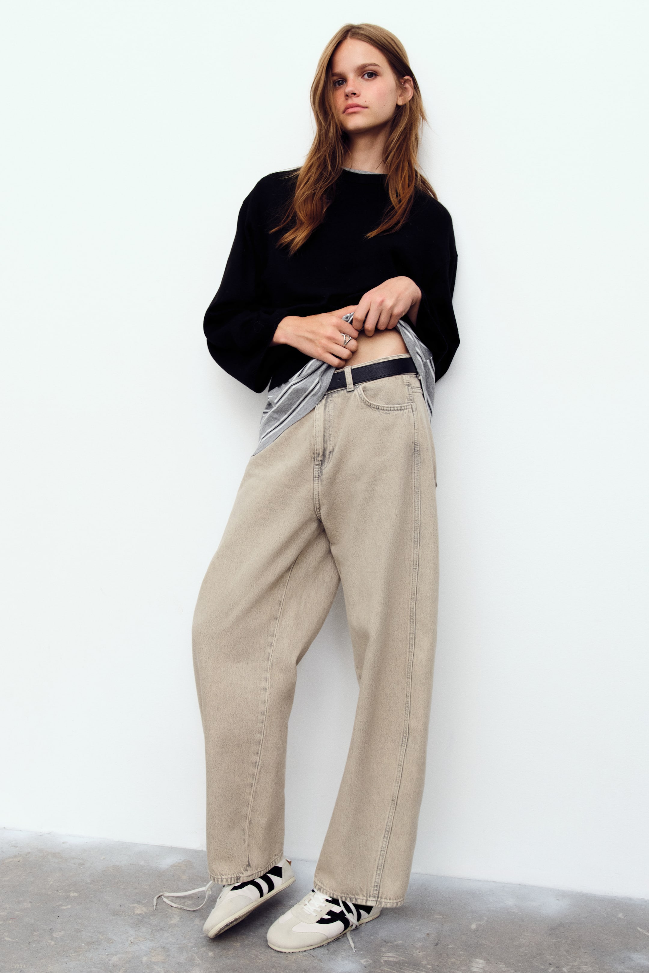 View larger image: The model is wearing a short black sweater in a soft knit, featuring a round neckline, dropped shoulders, and ribbed cuffs. The model is lifting the front hem of the sweater to reveal a grey striped shirt layered underneath, along with beige wide-leg jeans.