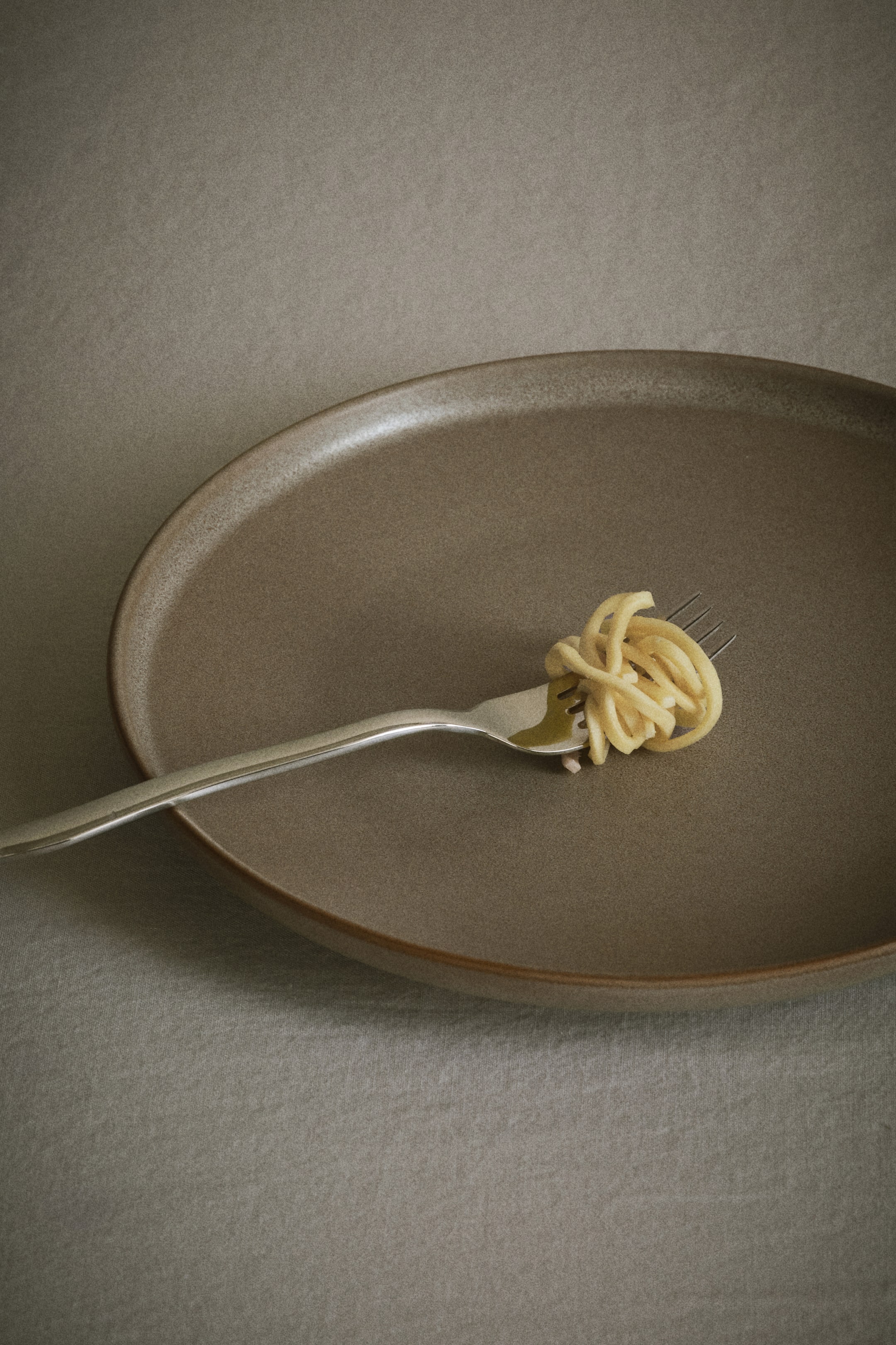 Voir une image plus grande: Une fourchette en argent repose sur une assiette gris-brun, ses dents tenant un petit paquet enroulé de spaghettis jaune pâle.
