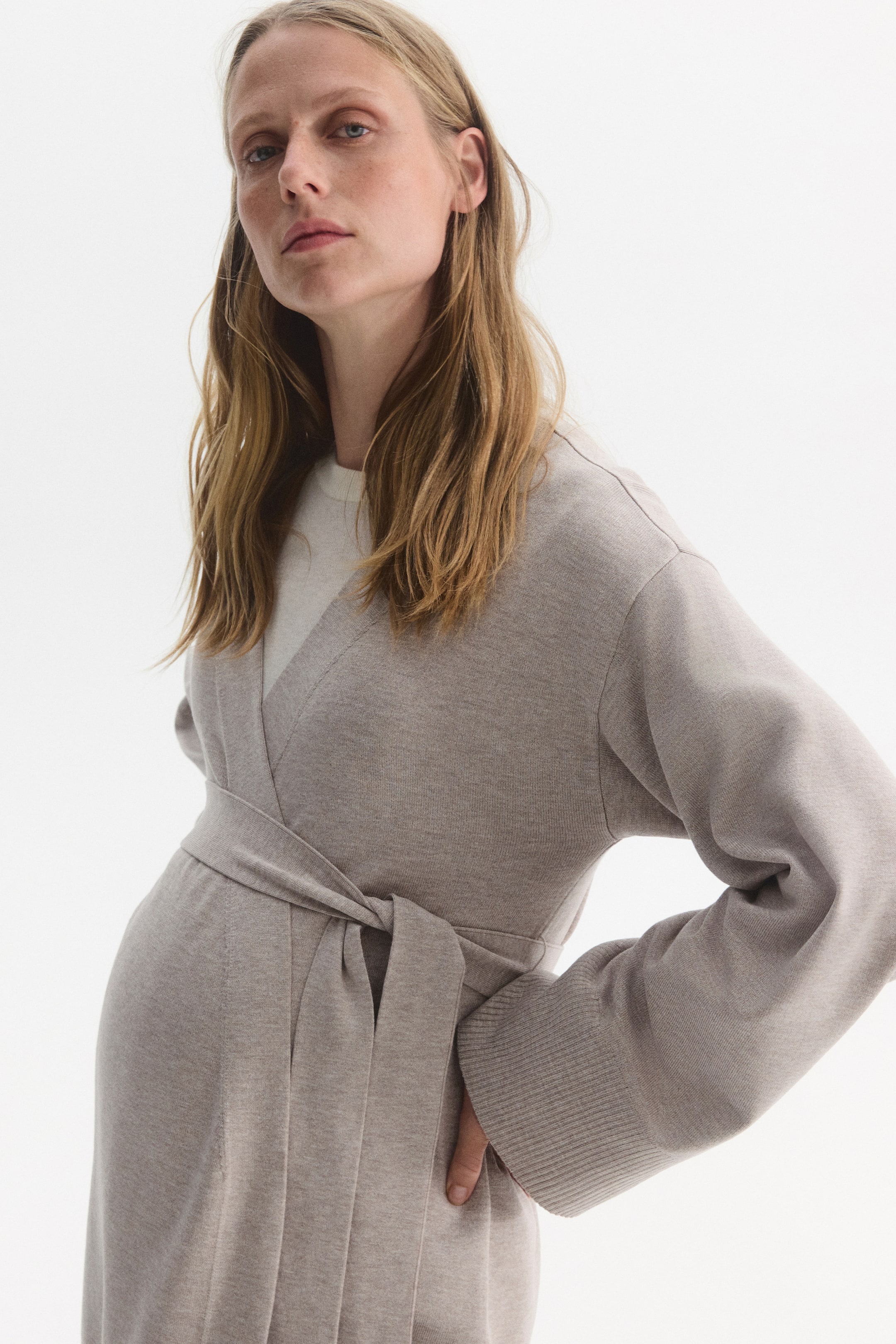 Größeres Bild anzeigen: Ein beigefarbener Cardigan aus weichem Strick wird vom Model über einem hellen Rundhals-Top getragen. Der Cardigan verfügt über überschnittene Schultern, lange Ärmel mit Rippbündchen und einen tiefen V-Ausschnitt, der durch die offene Front entsteht und mit einem farblich passenden Bindegürtel in der Taille geschlossen wird.