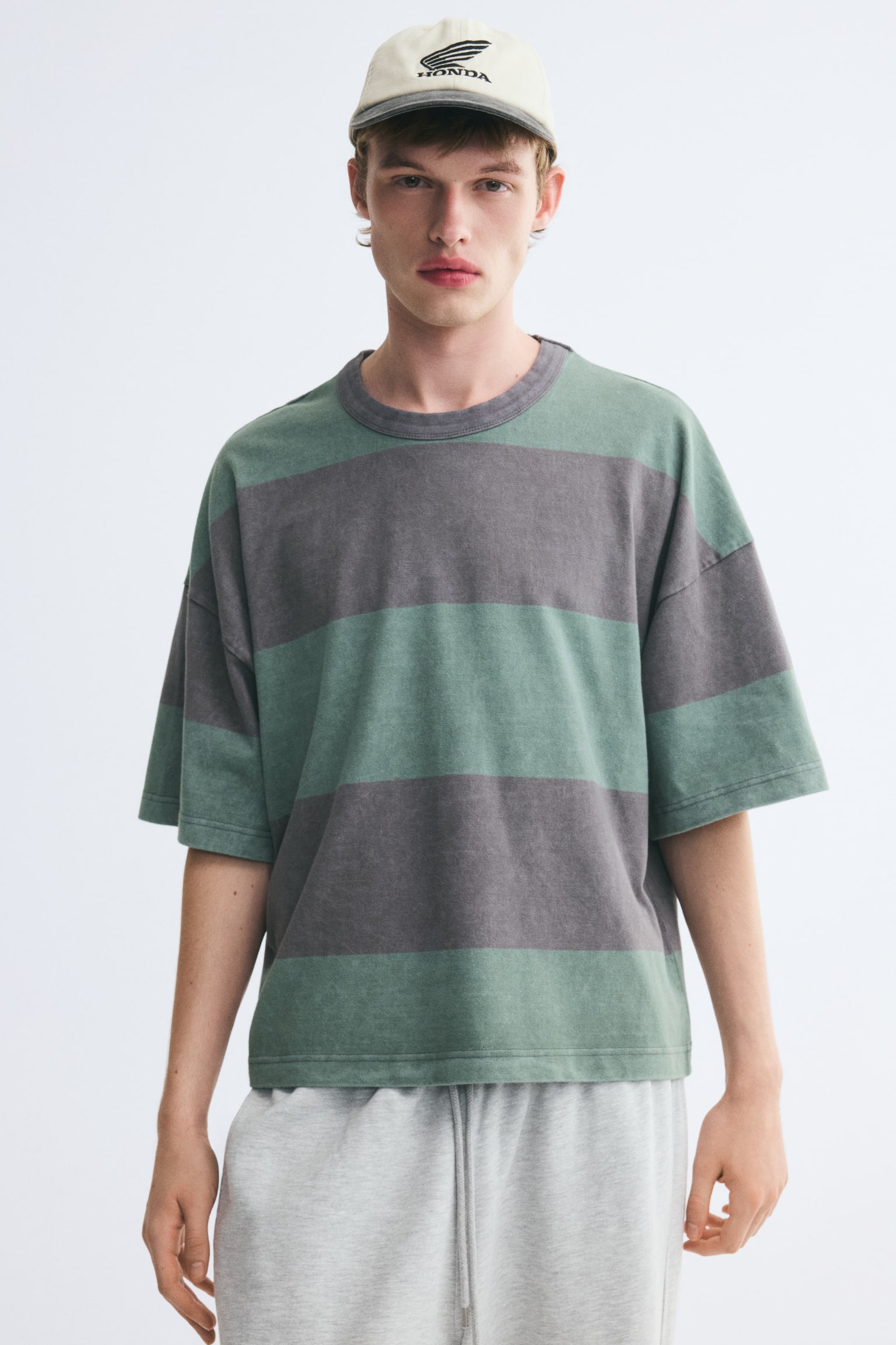 A boxy, oversized T-shirt with wide green and grey horizontal stripes is being worn by the model. The tee features a round, rib-trimmed grey neckline, dropped shoulders, and a straight-cut hem, and is paired with light grey drawstring sweatpants. - 1