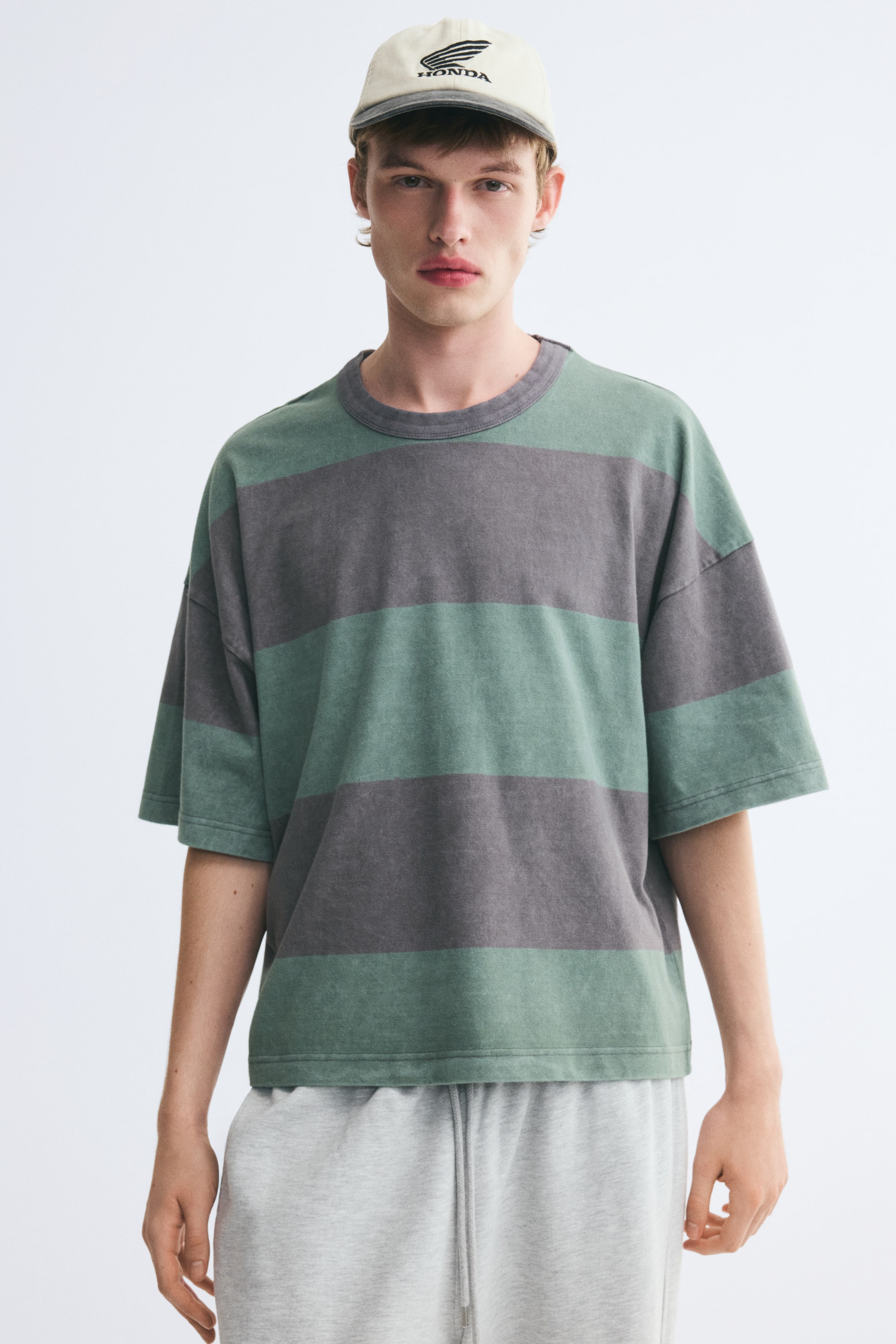 Vezi imaginea mai mare: Un tricou supradimensionat, cu croială boxy, cu dungi orizontale late verzi și gri, este purtat de model. Tricoul are un decolteu rotund gri, finisat cu bordură rib, umeri căzuți și tiv drept, și este asortat cu pantaloni de trening deschiși la culoare, gri, cu șnur.