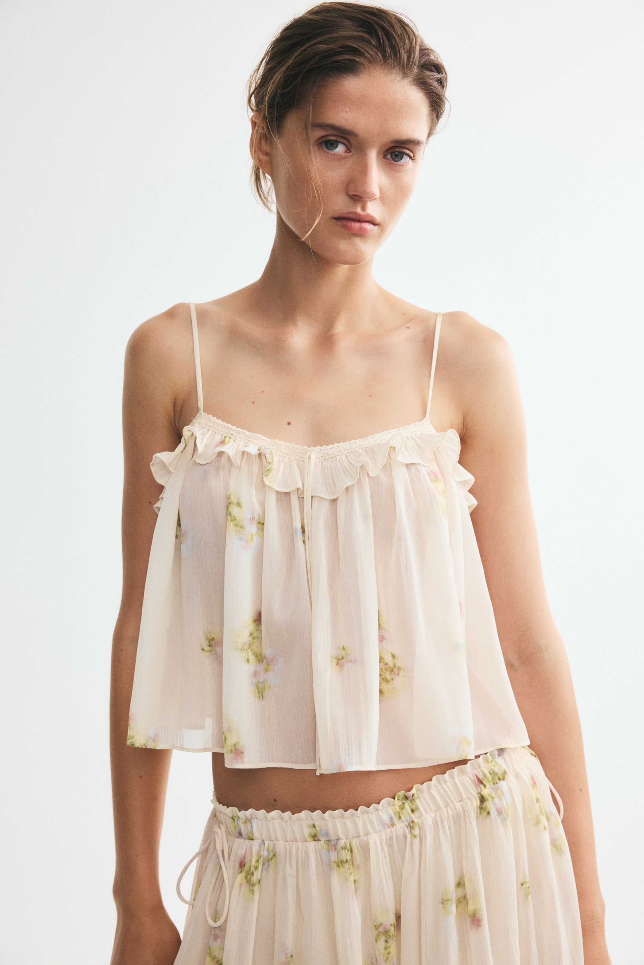 Grösseres Bild ansehen: Ein Cropped-Top aus gekräuseltem Chiffon mit zartem Blumenmuster wird vom Model getragen. Es verfügt über einen mit Spitze besetzten V-Ausschnitt mit rundum Rüschenbesatz, Spaghettibänder vorne, schmale, verstellbare Träger und einen sanft gerundeten Saum; dazu trägt das Model die passende Shorts.