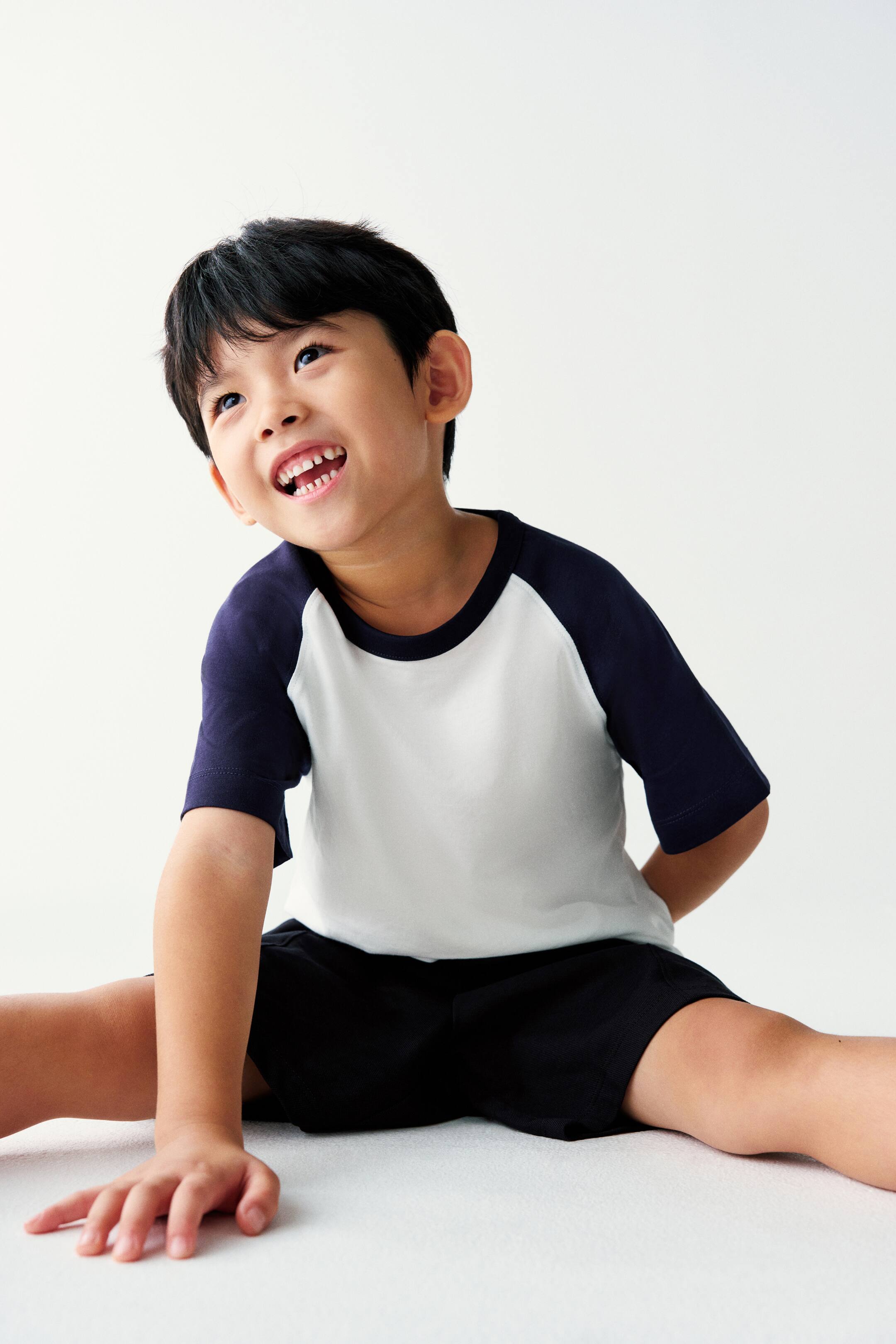 View larger image: The model is wearing a short-sleeved raglan T-shirt with a white body, navy blue sleeves, and a navy crew neck, paired with simple black shorts.