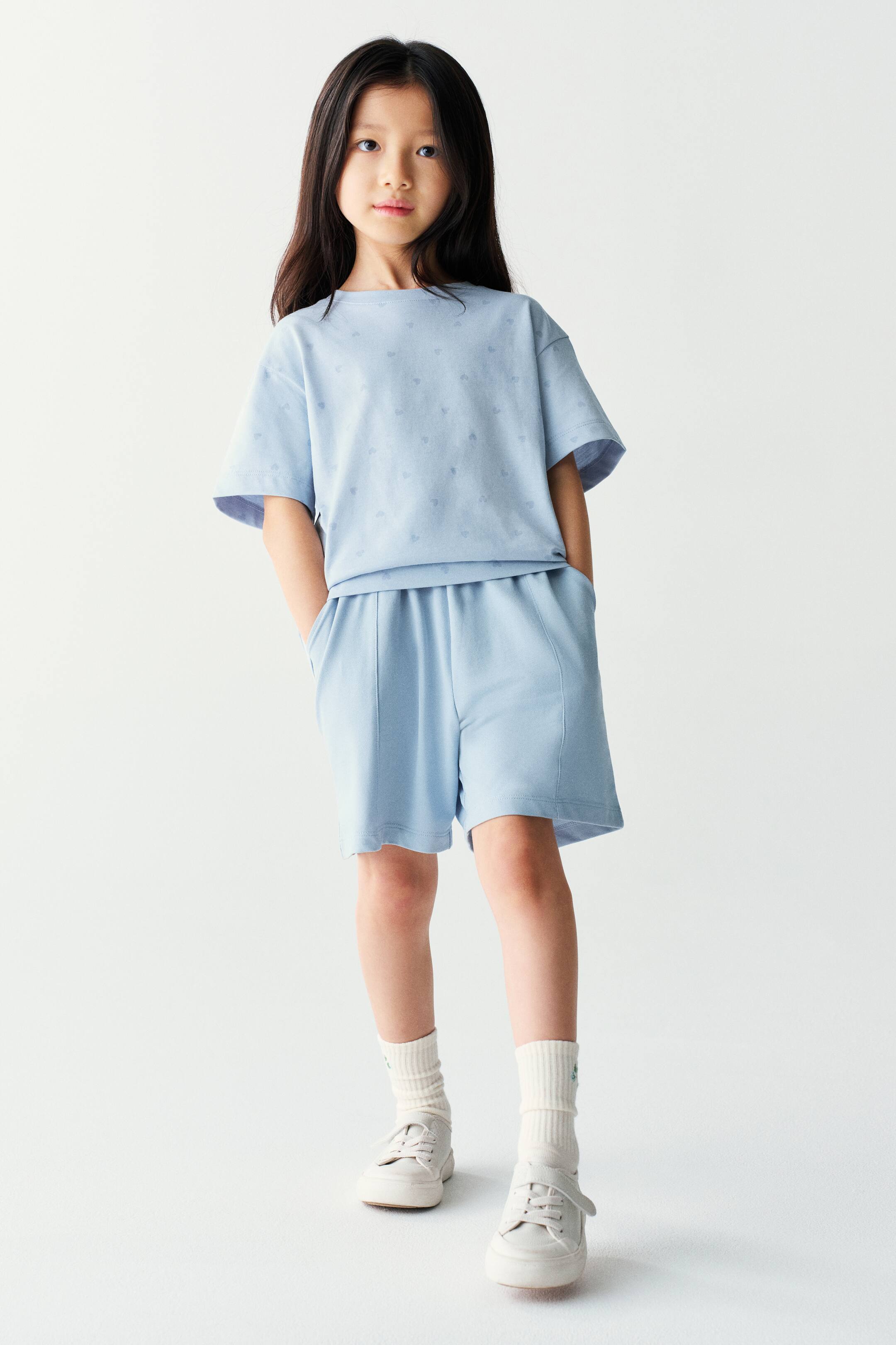 View larger image: The model is wearing a light blue matching set consisting of a short-sleeved t-shirt with a subtle print and coordinating shorts. The shorts feature front seam details and side pockets, with the model placing their hands in the pockets.