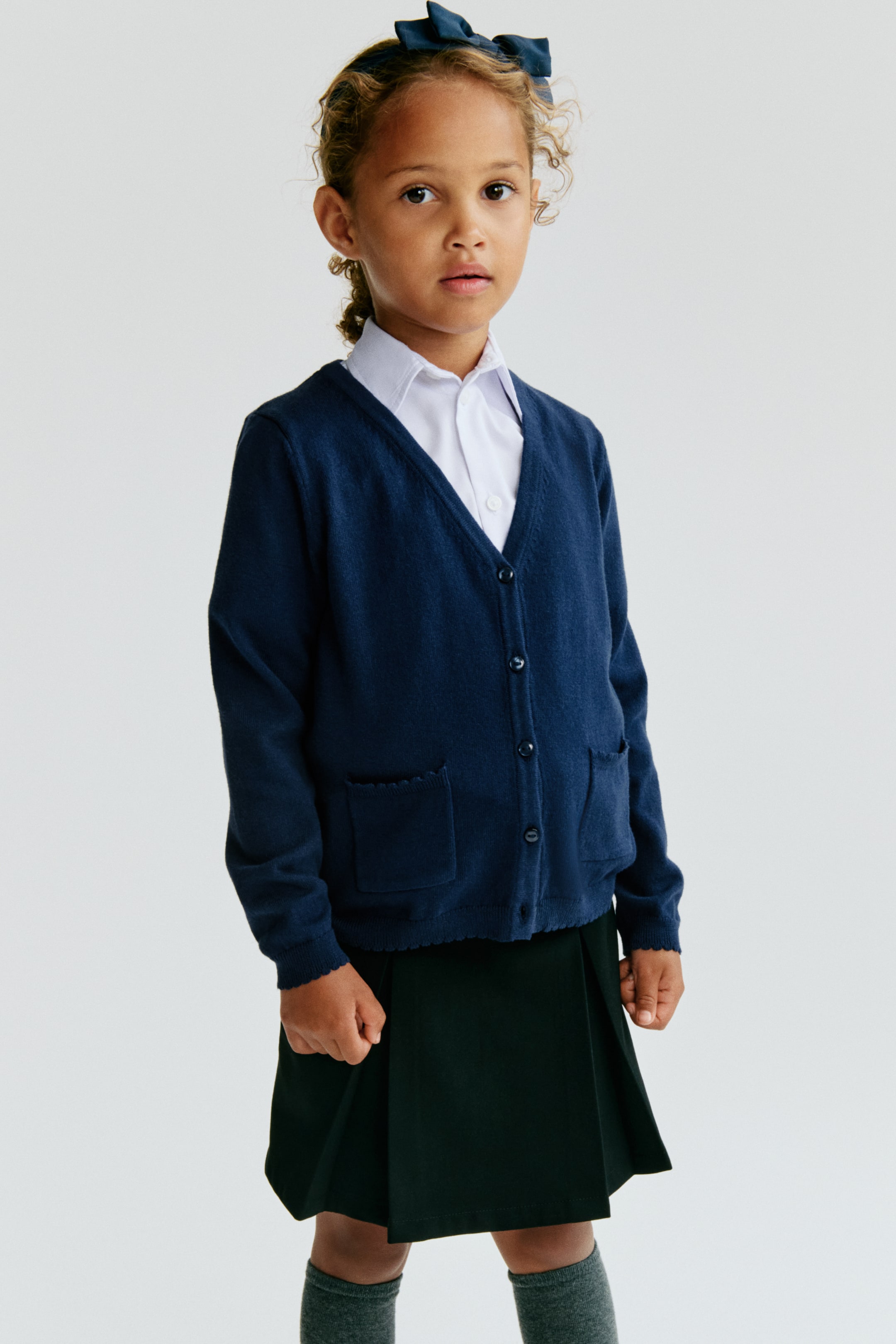 Voir une image plus grande: Un cardigan bleu marine présente un design à col en V et une fermeture à boutons sur le devant avec des boutons foncés. Il est doté de deux poches plaquées à l'avant avec des bords festonnés, ainsi que de poignets et d'un ourlet côtelés, et est porté par-dessus une chemise à col blanc.