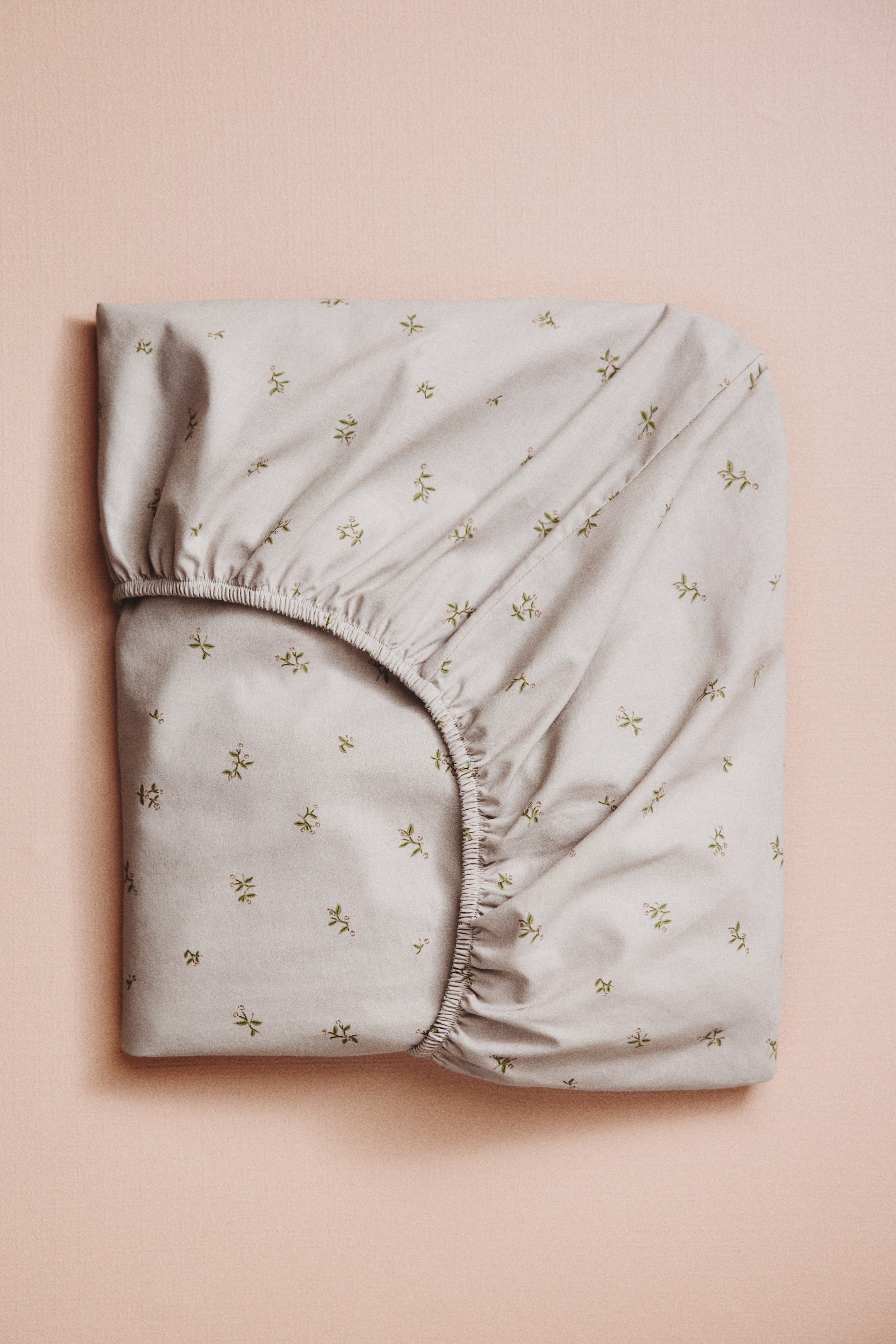 Agrandir l'image: Un drap-housse en tissage de coton gris clair, agrémenté d'un motif floral vert dispersé, est plié avec un coin élastiqué visible, révélant son design ajusté.