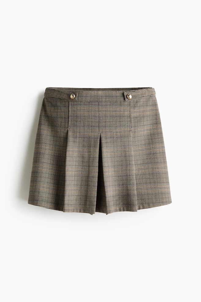 A front view of a brown and beige plaid mini skirt is showing a structured waistband with two decorative side tabs adorned with round, gold-tone buttons. The skirt is featuring deep box pleats along the front, creating a structured, flared silhouette. - 1