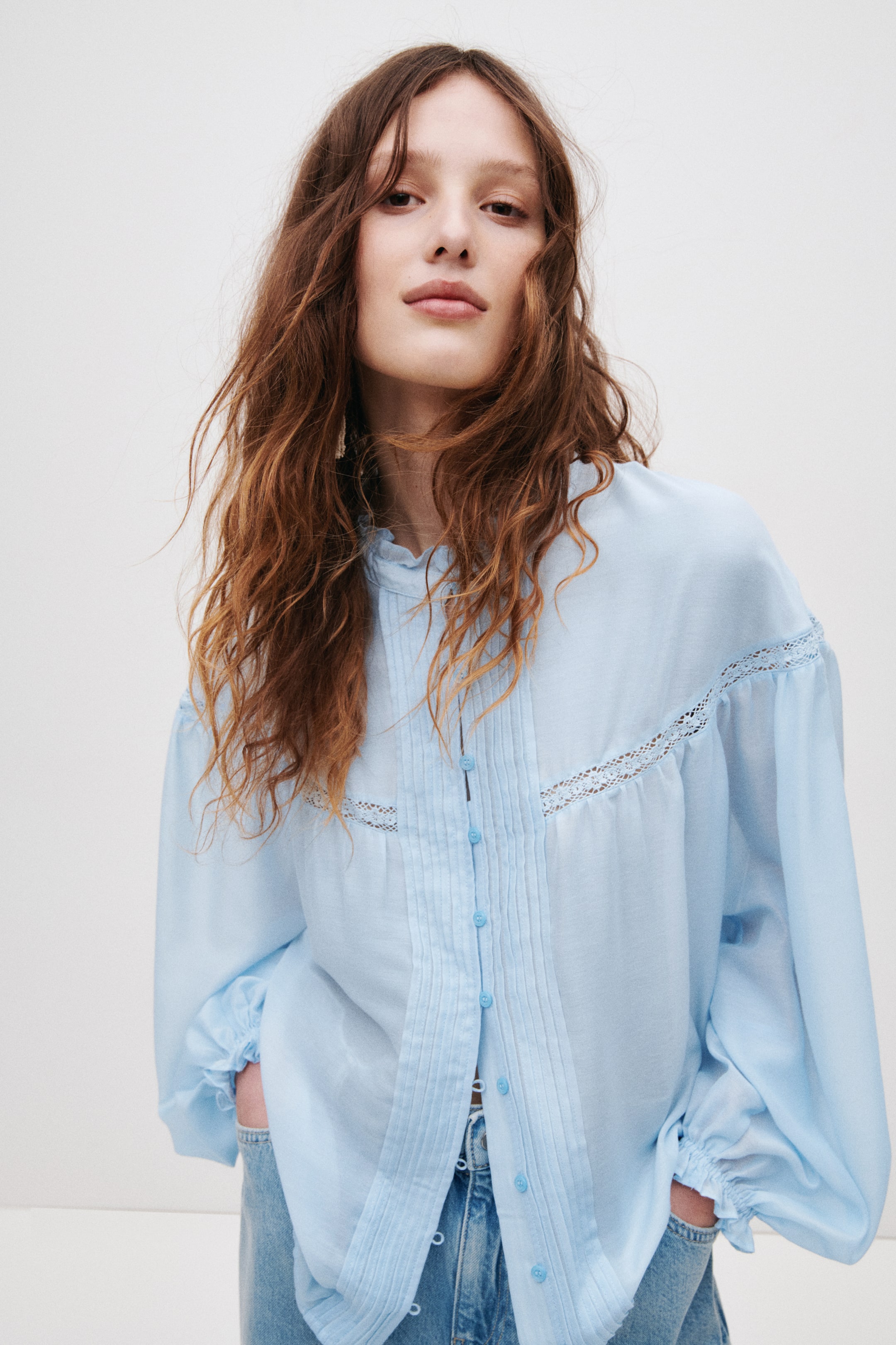 Grotere afbeelding bekijken: Een lichtblauwe blouse met een gerimpelde opstaande kraag en een volledige knoopsluiting met bijpassende knopen. Verticale plooien sieren de voorkant, met een horizontale kanten bies die over de borst en schouders loopt, en lange volumineuze mouwen eindigen in elastische manchetten. De blouse is gedeeltelijk ingestopt in blauwe jeans.