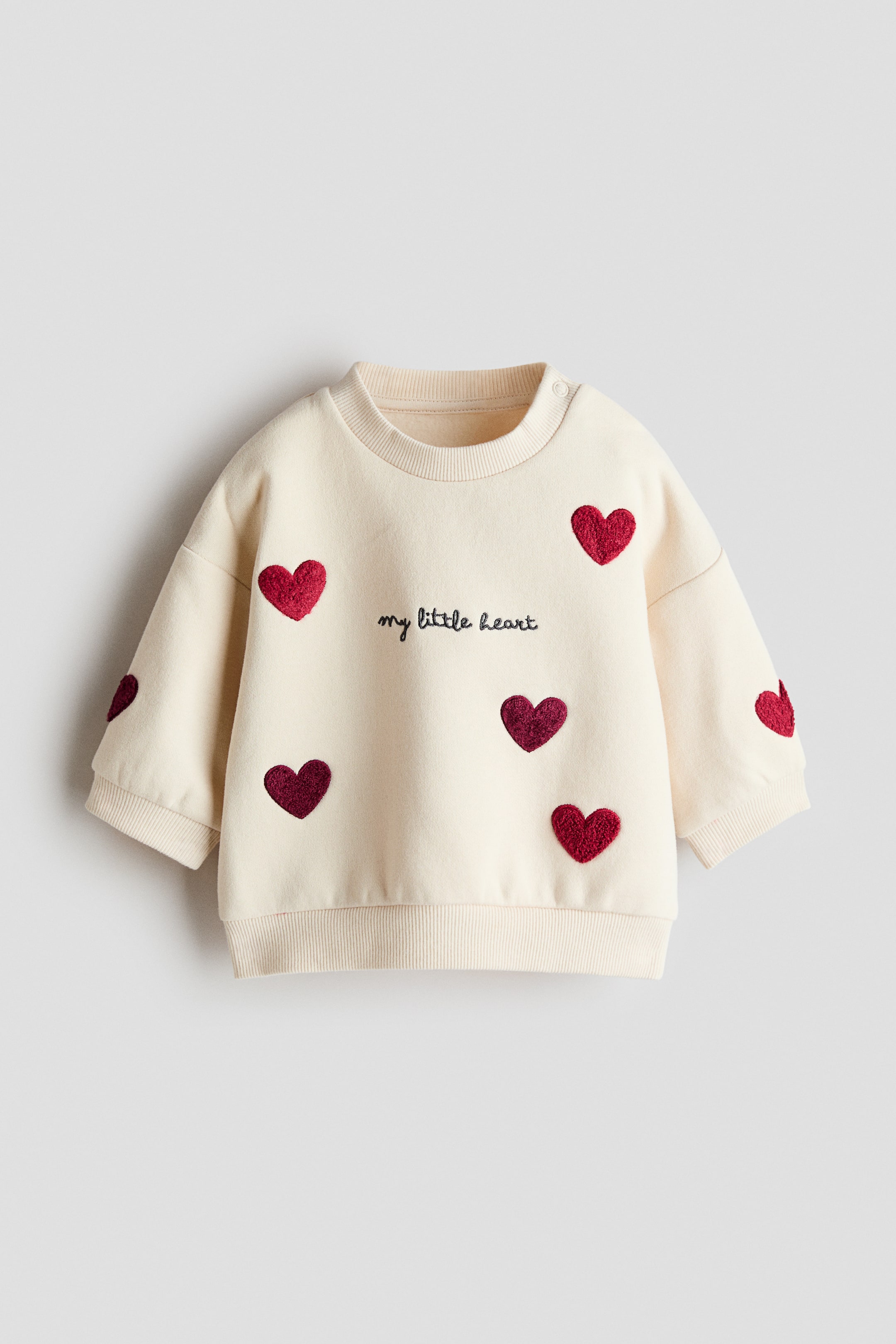 View larger image: A cream-coloured sweatshirt is showcased from the front, boasting a ribbed crew neck with two snap buttons on the left shoulder. It is embellished with black embroidered text reading "my little heart" and numerous red and burgundy heart appliqués, finished with ribbed cuffs and a ribbed hem.
