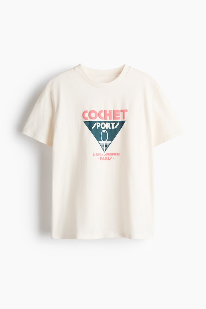 A cream T-shirt in printed cotton jersey is presented from the front, featuring a round, rib-trimmed neckline, gently dropped shoulders, and a straight-cut hem. A graphic print is centered on its front, displaying "COCHET" in pink above a dark teal triangle containing "SPORT" and a tennis racket illustration, with "21 RUE DE LA P�PINI�RE PARIS" in pink below. - 1