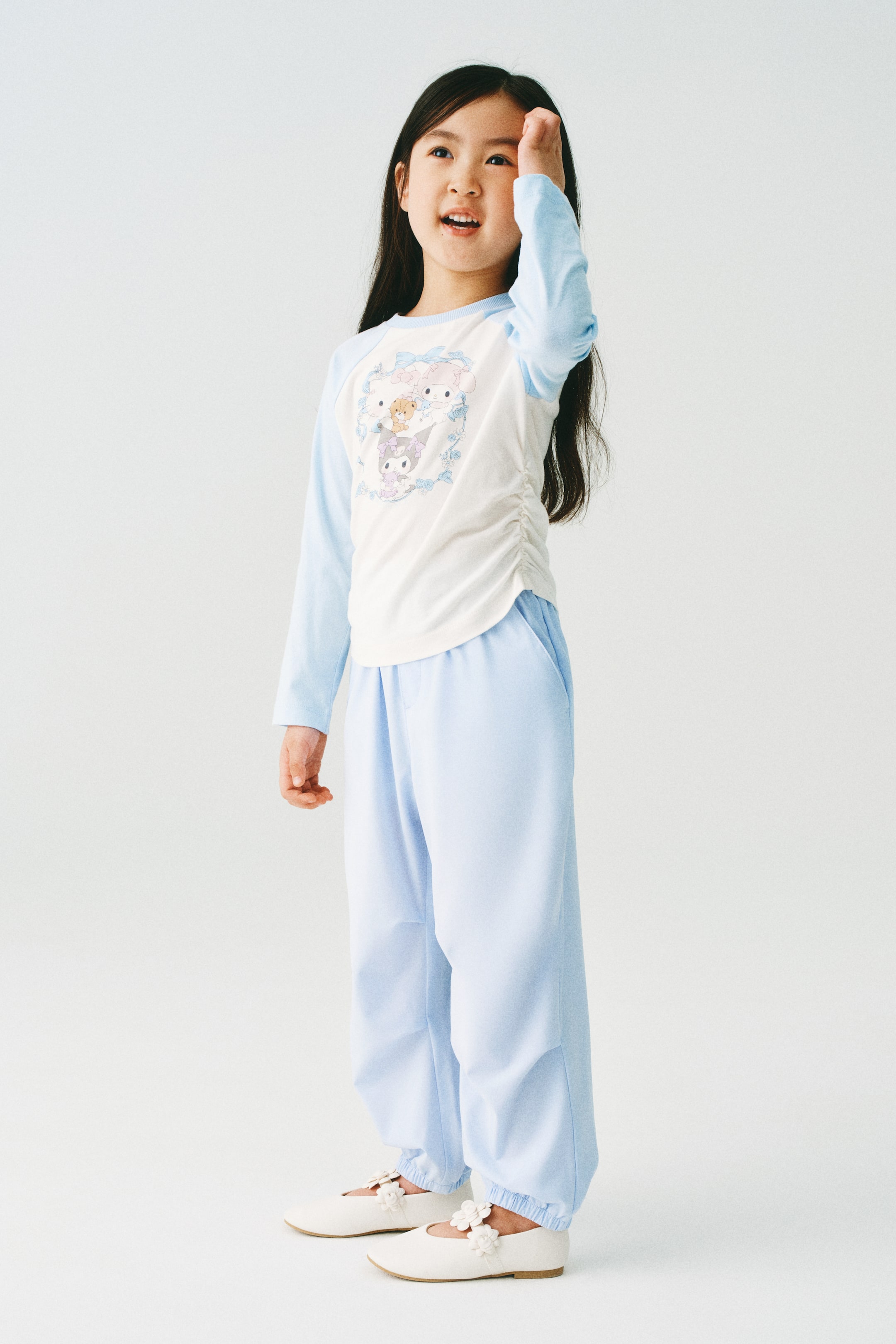 拡大画像を表示: モデルが着用しているのは、織物素材のライトブルーのジョガーパンツです。ウエストはゴム仕様で、斜めのサイドポケットと裾にはカバー付きのゴムが施されています。モデルはまた、ライトブルーとホワイトの長袖シャツと、ホワイトのメリージェーンシューズも着用しています。