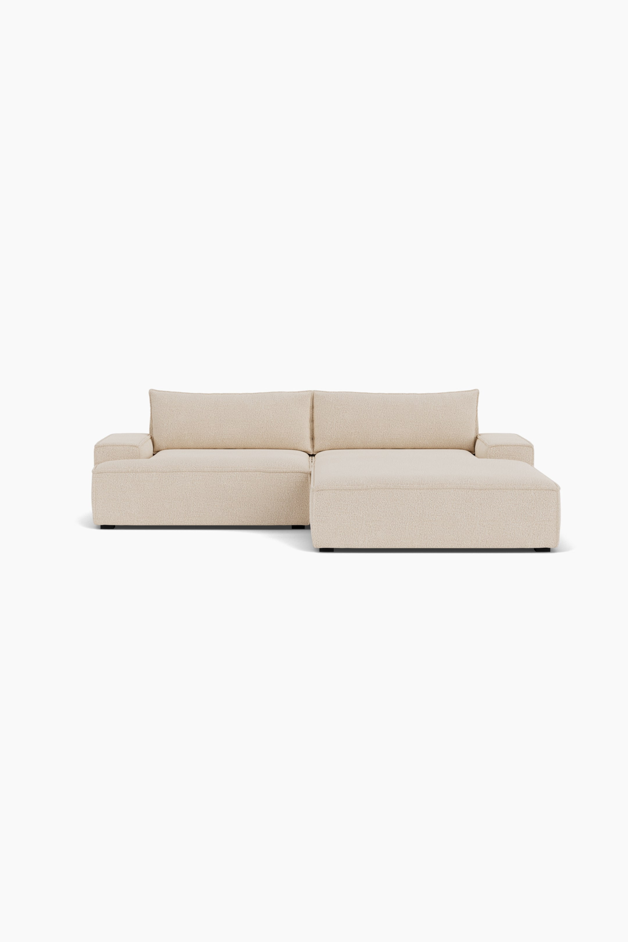 Grotere afbeelding bekijken: Een crèmekleurige modulaire hoekbank staat recht naar voren gericht en laat de gestructureerde beige bekleding en het lage profielontwerp zien. De bank is opgesteld met een standaard zitgedeelte links en een verlengde chaise lounge rechts, met dikke, blokvormige armleuningen en twee zachte rugkussens.