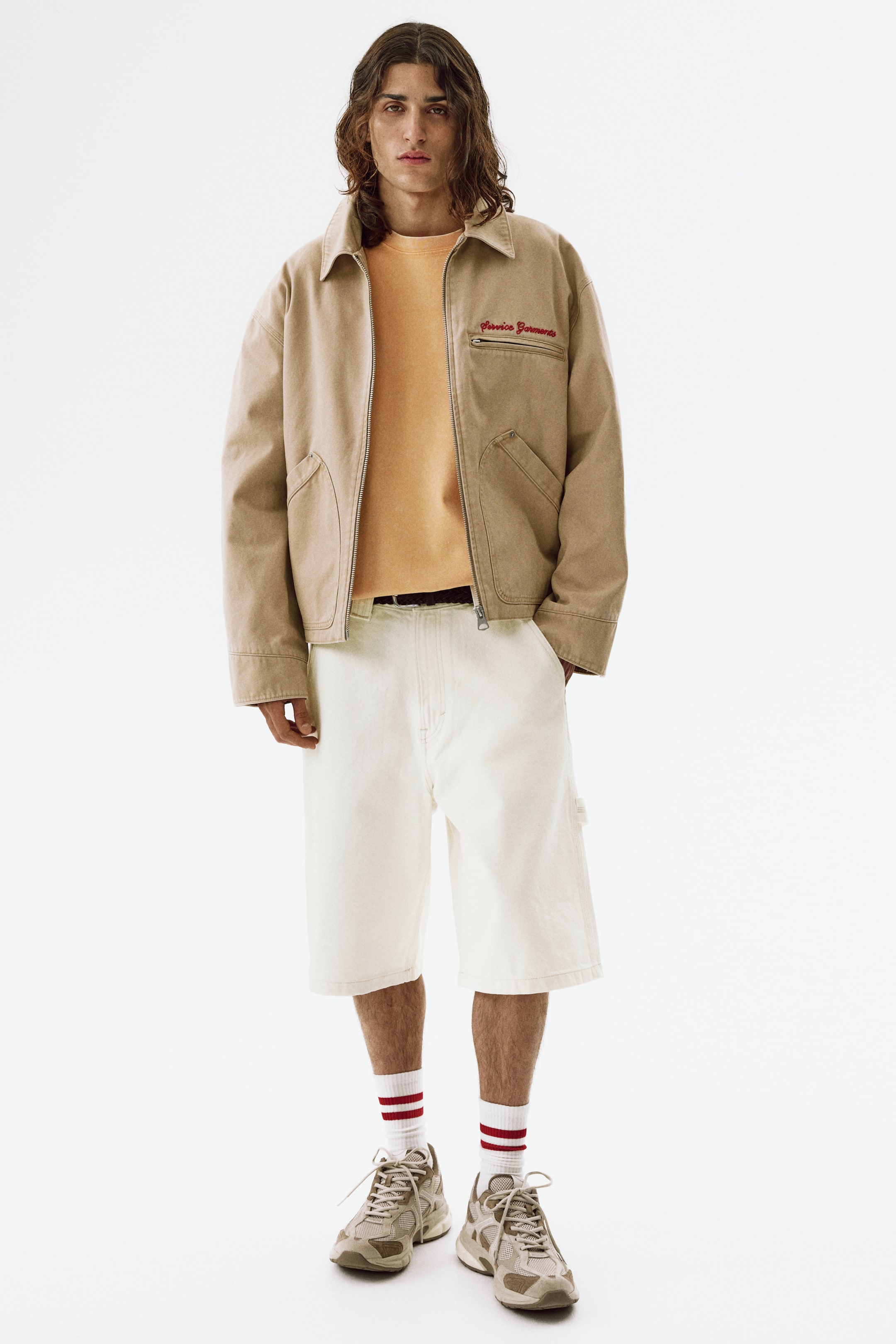 View larger image: Presenting a tan workwear jacket, this piece features a pointed collar and a full-zip front closure. It showcases two lower patch pockets and a zippered chest pocket adorned with red "Service Garments" embroidery, styled open over a base layer.