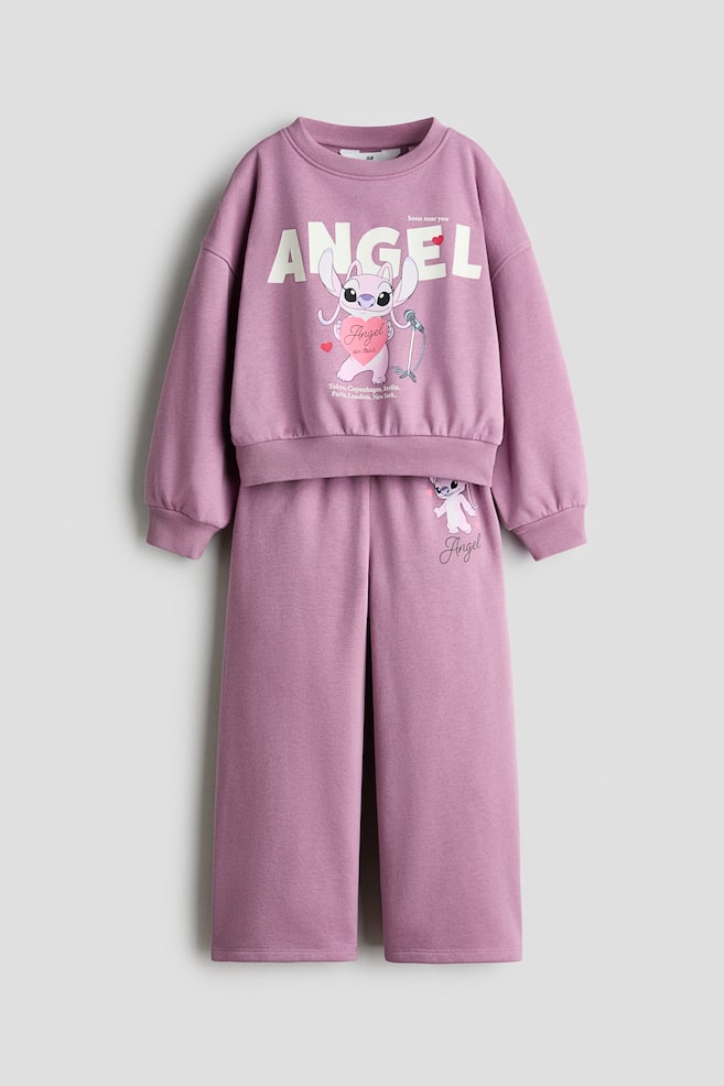 Ein zweiteiliges lila Loungewear-Set wird von vorne gezeigt, bestehend aus einem verkürzten Sweatshirt mit Rundhalsausschnitt und einer passenden weiten Hose. Auf dem Sweatshirt ist eine groÃe Grafik der Figur Angel unter dem in kräftigen weiÃen Buchstaben gedruckten Wort âANGELâ zu sehen; abgerundet durch gerippte Bündchen und einen gerippten Saum. Die Hose zeigt eine kleinere passende Figuren-Grafik auf dem oberen linken Oberschenkel. - 1