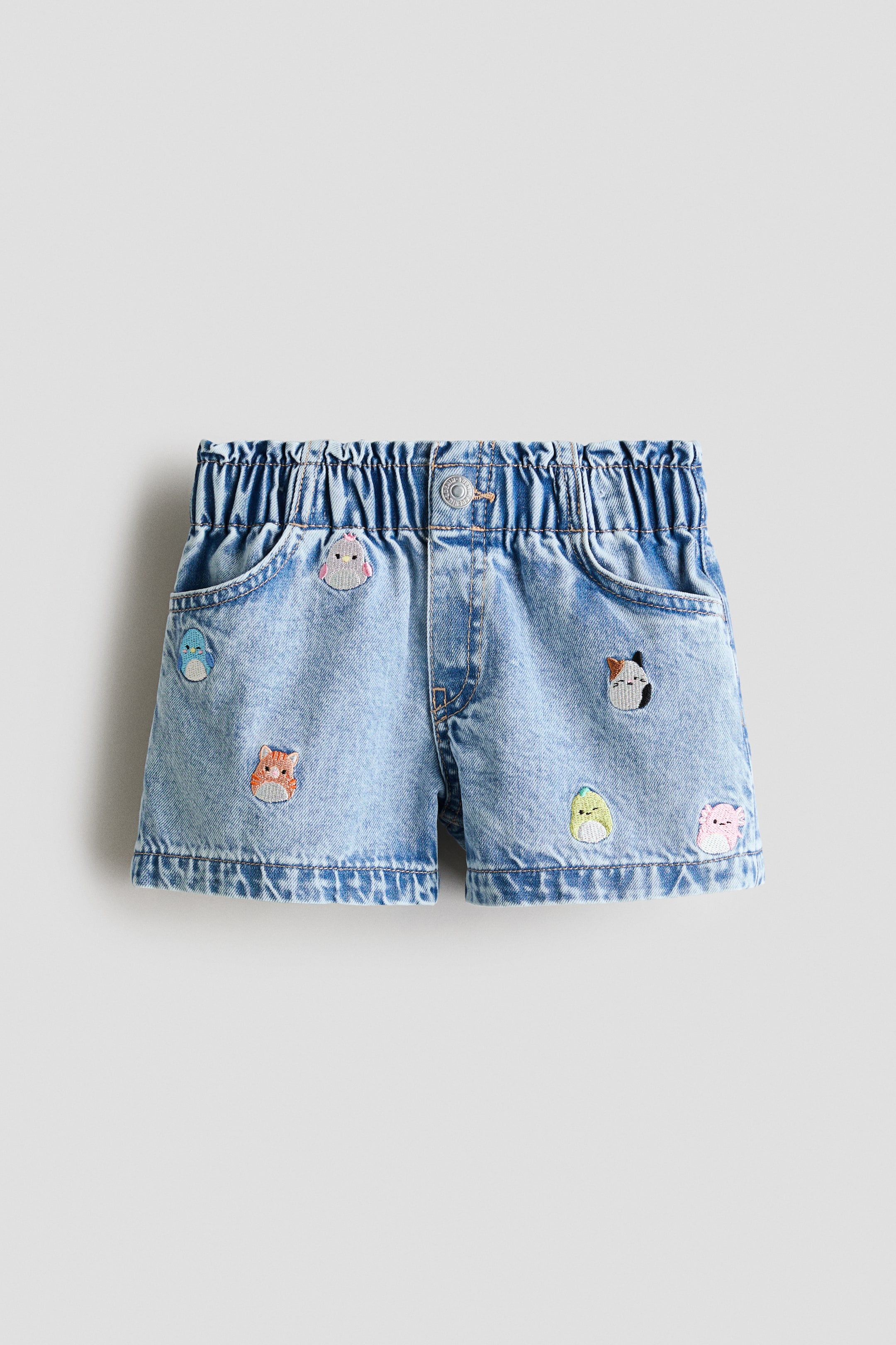View larger image: Denim shorts are showcased from the front, featuring a light wash and a relaxed fit. They are crafted with an elasticated paper bag waist, a buttoned faux fly, and diagonal front pockets, while various embroidered characters adorn the surface.