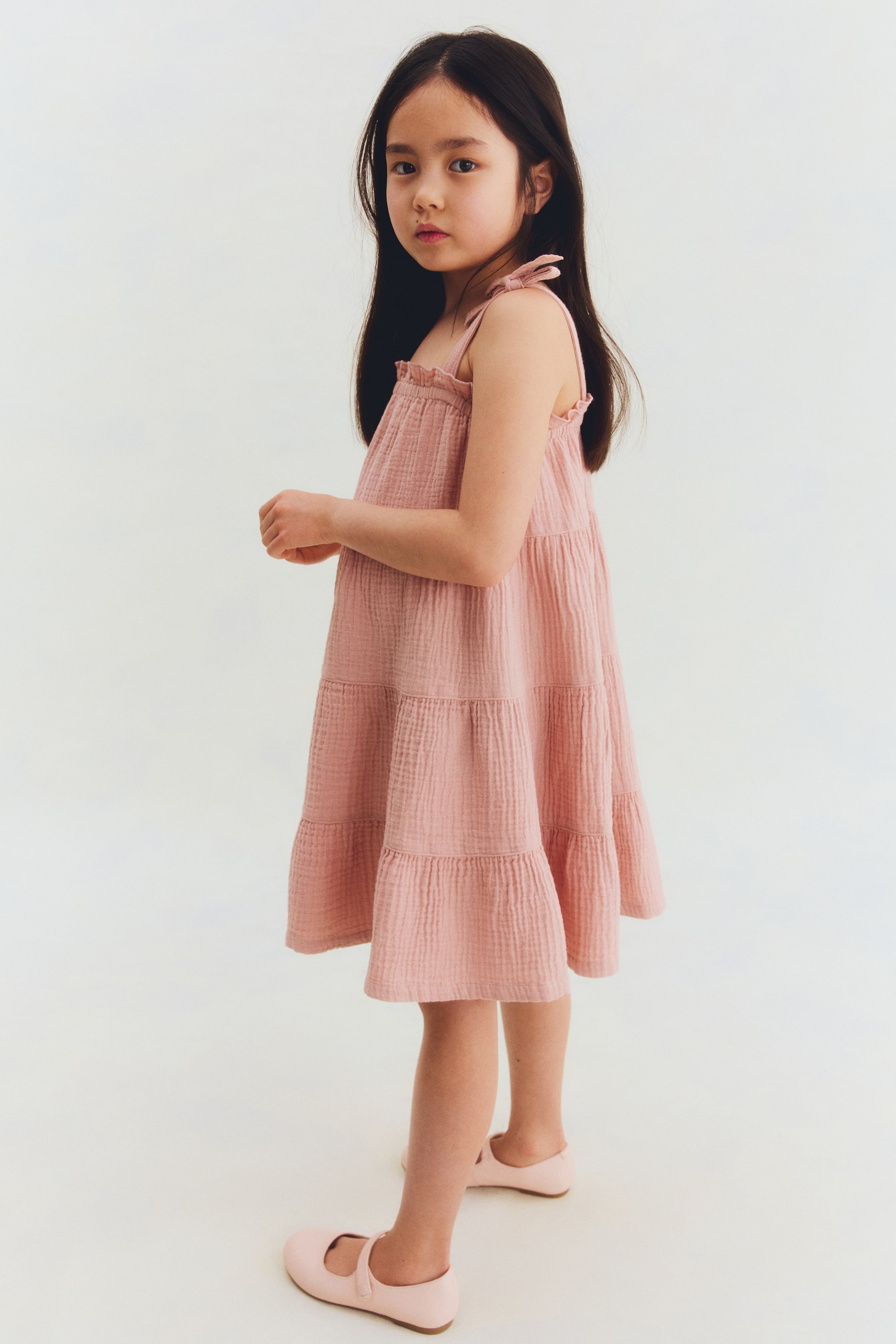 View larger image: A pink tiered dress showcases a loose silhouette from a slightly angled front view. It features delicate tie-bow shoulder straps, a gathered neckline, and a crinkled texture, with multiple horizontal tiers creating a relaxed, flowy shape.