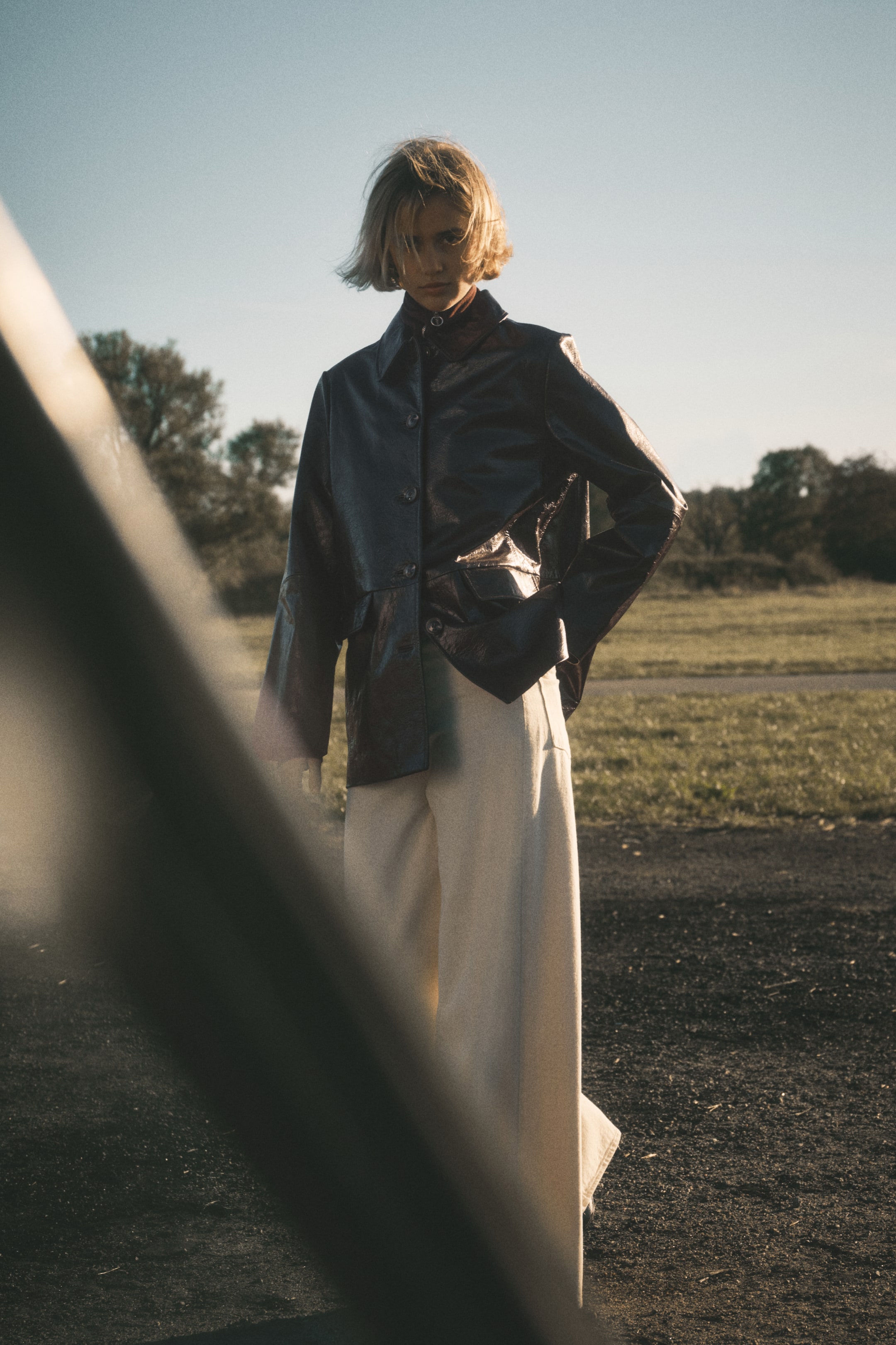 Ingrandisci l'immagine: Una giacca scura e lucida presenta un colletto aperto e una chiusura frontale con bottoni, stratificata su un top scuro. Si abbina con pantaloni a gamba larga di colore chiaro, il tutto visibile da un leggero angolo frontale sinistro.