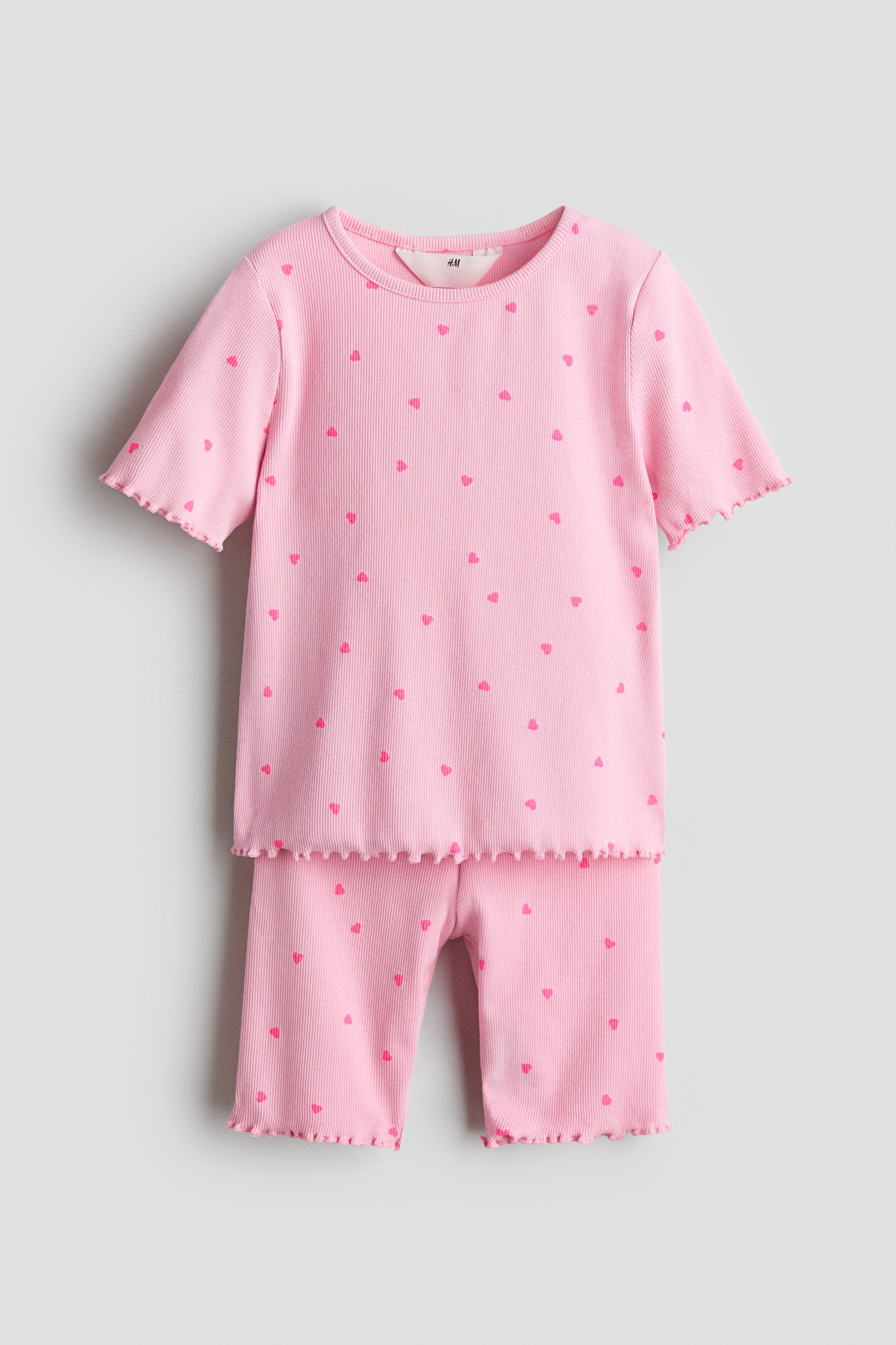 拡大画像を表示: ピンクのリブパジャマセットは、正面を向いており、ショートスリーブのトップとカプリ丈のボトムス全体に小さな濃いピンクのハートが散りばめられています。トップはラウンドネックラインで、袖口と裾にレタスエッジのトリムが施されており、ボトムスの脚開口部のレタスエッジトリムと一致しています。