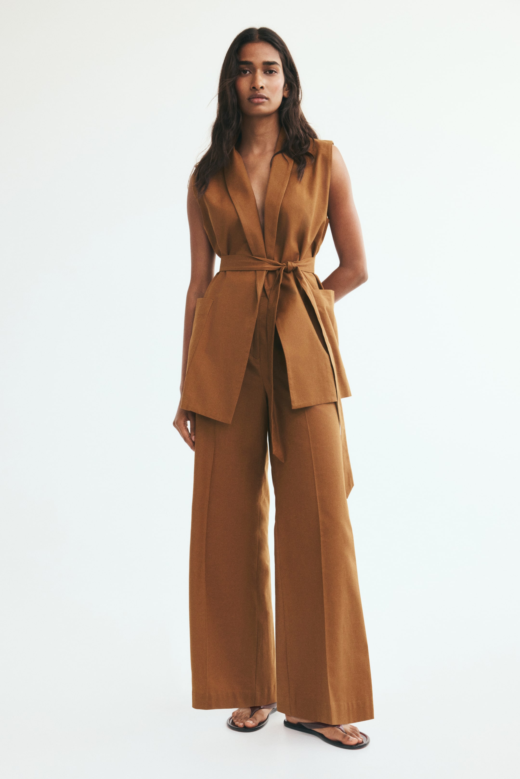 View larger image: The model is wearing brown wide-leg trousers with front creases. They are paired with a matching sleeveless belted waistcoat in a textured linen-blend fabric.