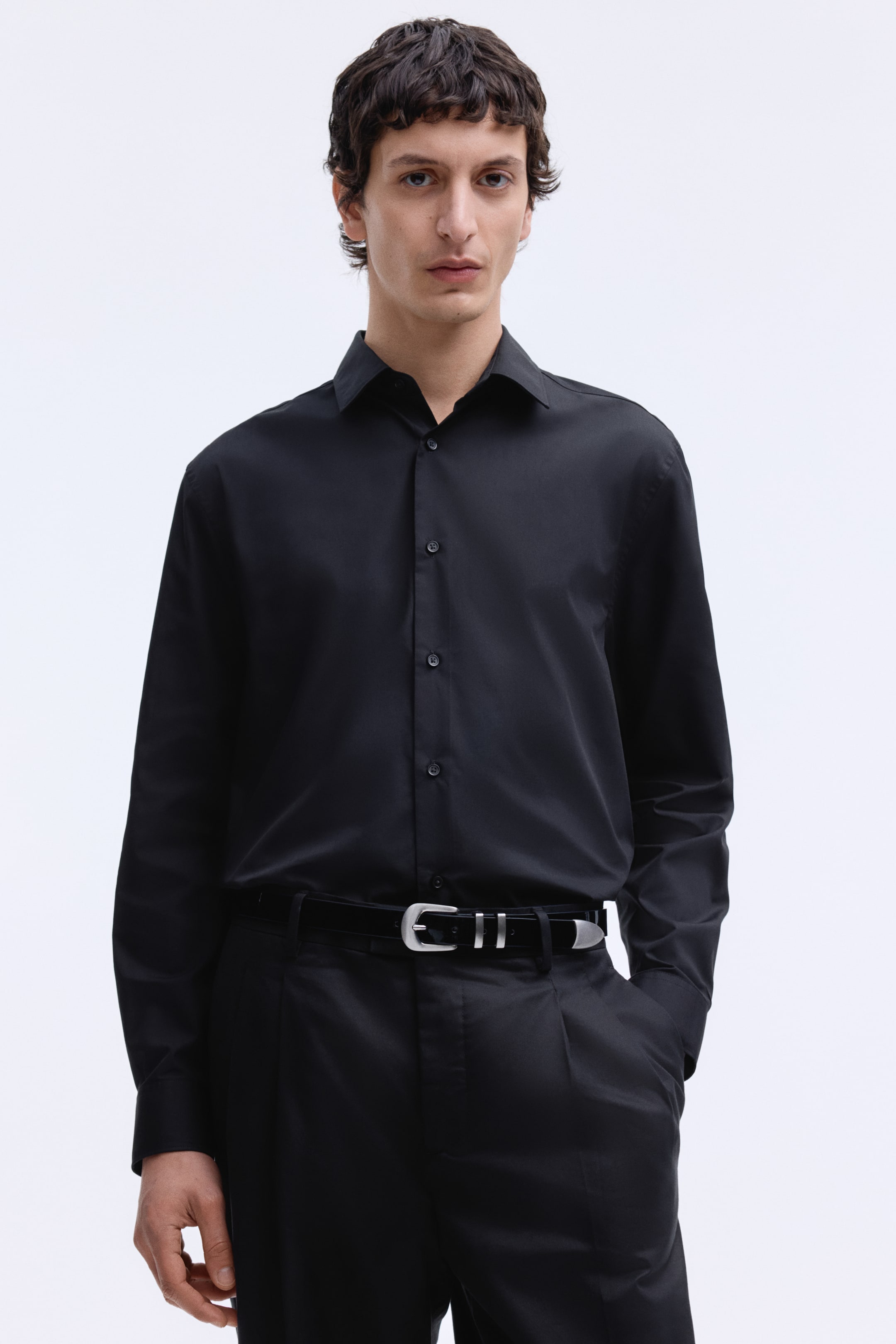 View larger image: A black long-sleeved shirt featuring a pointed collar and a full-button placket with dark buttons, showcasing a smooth fabric texture. It's neatly tucked into black trousers and secured by a black belt with a silver buckle.