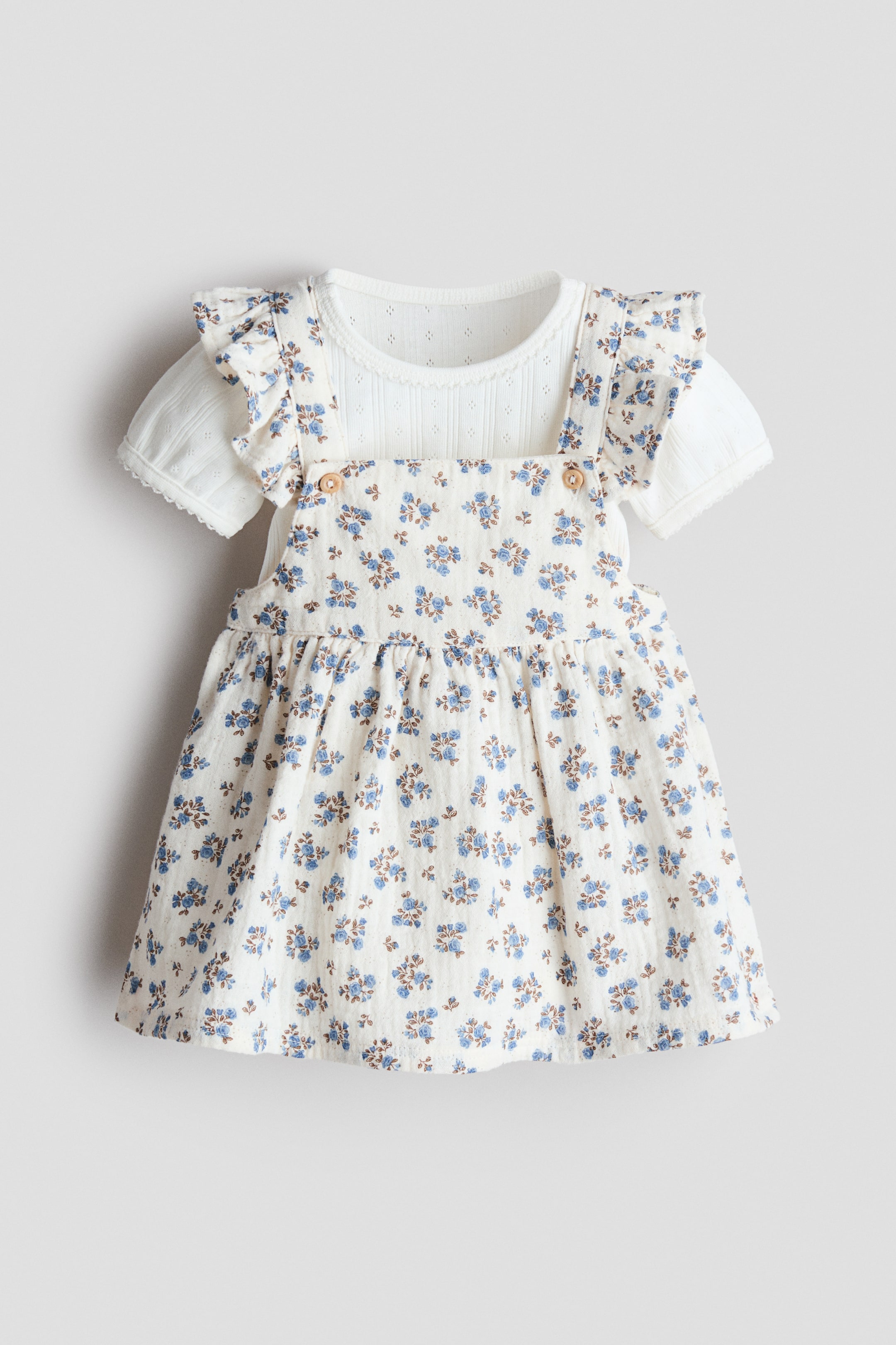 View larger image: A baby's floral pinafore dress set features a cream-coloured pinafore dress with an all-over blue floral print and shoulder straps fastening with small brown buttons at the chest. A white short-sleeved top layers underneath, showcasing a textured knit fabric with eyelet details and ruffled sleeves with scalloped lace trim.