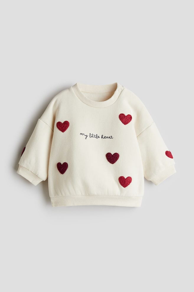 A motif-detail sweatshirt is presented from the front, in cream, featuring a ribbed crew neck, ribbed cuffs, and a ribbed hem. Its front is adorned with scattered heart-shaped appliques in various shades of red, some appearing glittery, and black embroidered text reading 'my little heart' is positioned centrally on the chest. A snap button closure is visible on the right shoulder. - 1