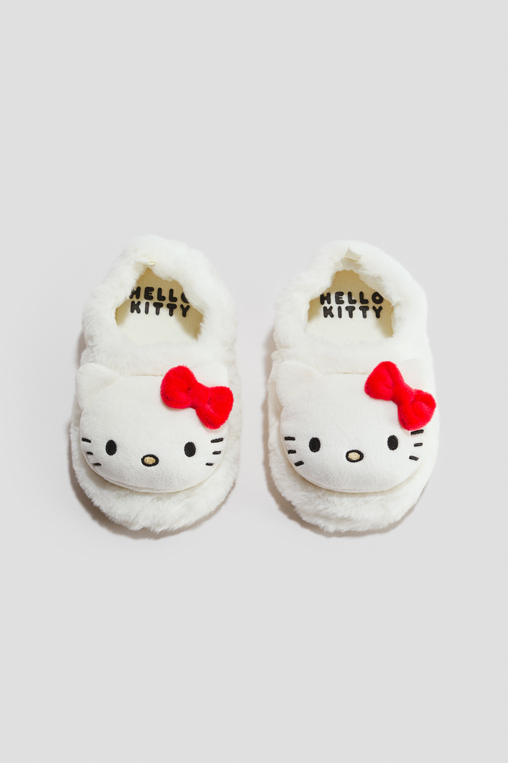 Agrandir l'image: Chaussons doux. Une paire de chaussons blancs et moelleux est présentée de face, chaque chausson arborant un visage de personnage blanc avec des yeux noirs, un nez jaune et des moustaches. Un nœud rouge orne le côté supérieur droit de chaque visage, et la semelle intérieure de couleur claire affiche les mots "HELLO KITTY" en noir.