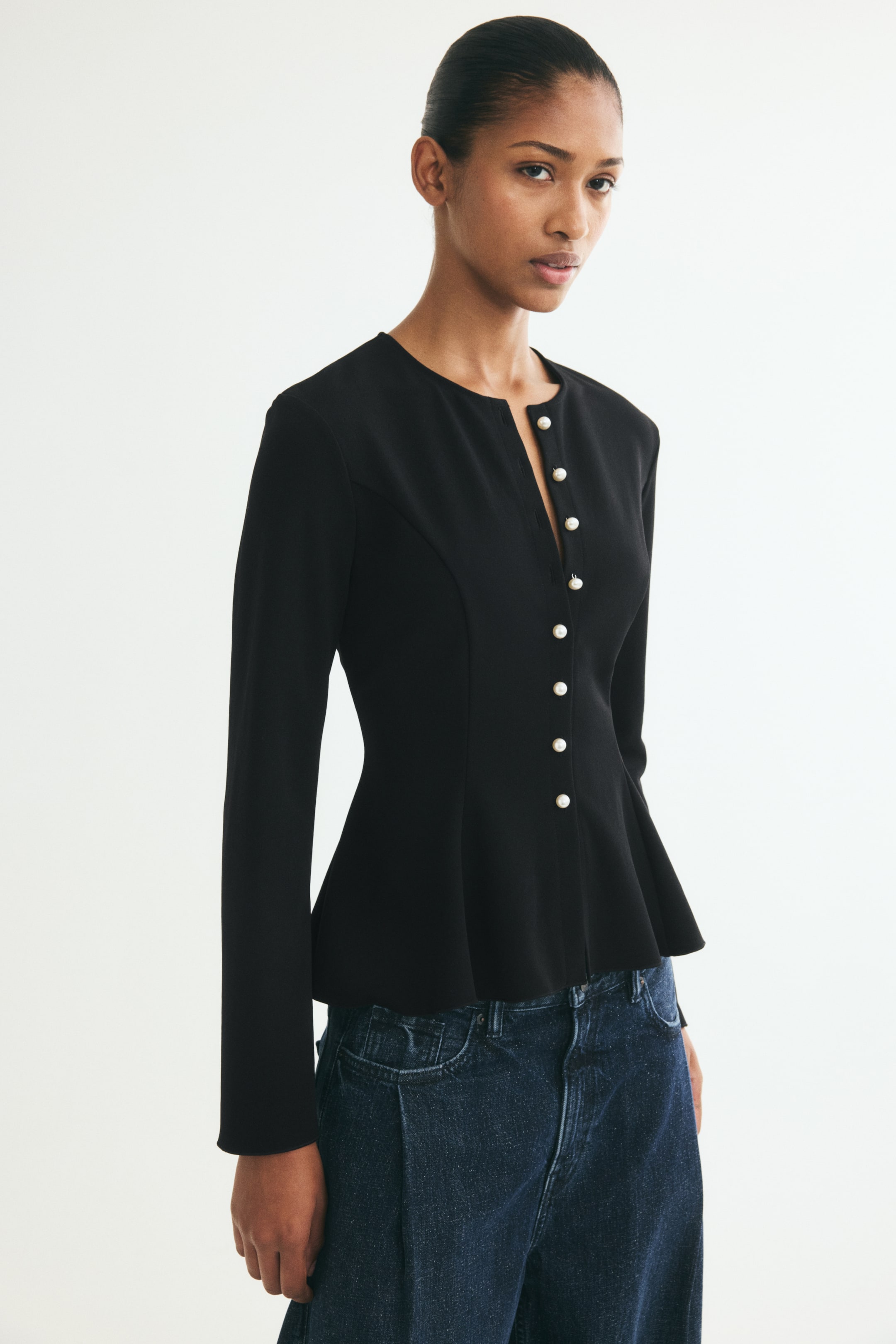 Xem ảnh lớn hơn: Người mẫu đang mặc một chiếc áo peplum tay dài màu đen, có phom ôm sát, cổ tròn không cổ với đường xẻ nhỏ phía trước, và hàng cúc trắng giống ngọc trai. Áo được phối cùng quần jeans denim xanh đậm.