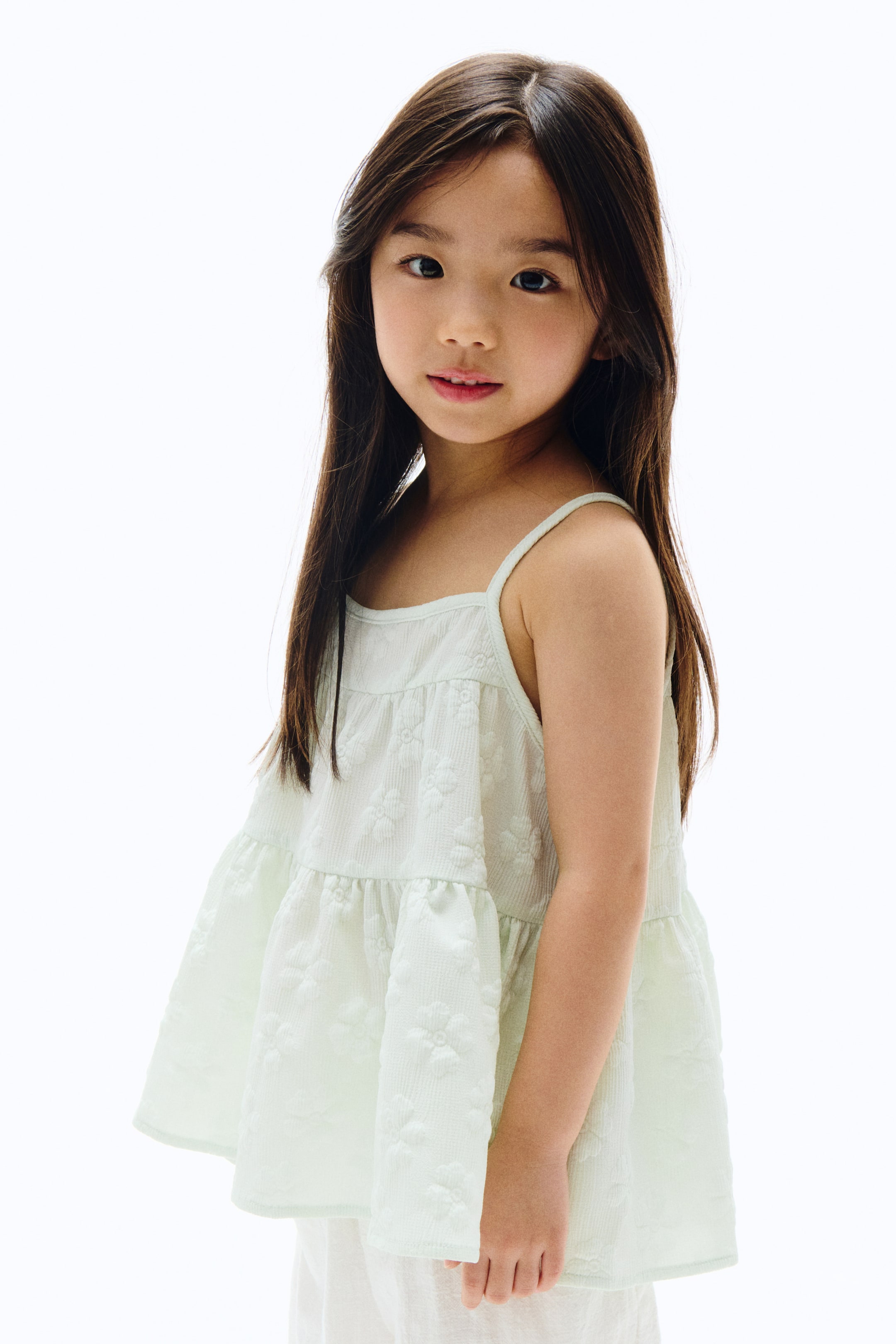 拡大画像を表示: ライトグリーンのトップは、細いスパゲッティストラップとストレートネックラインが特徴です。全体にテクスチャードフローラルパターンを披露し、段々になったゆったりとしたデザインが少し横向きに紹介されています。