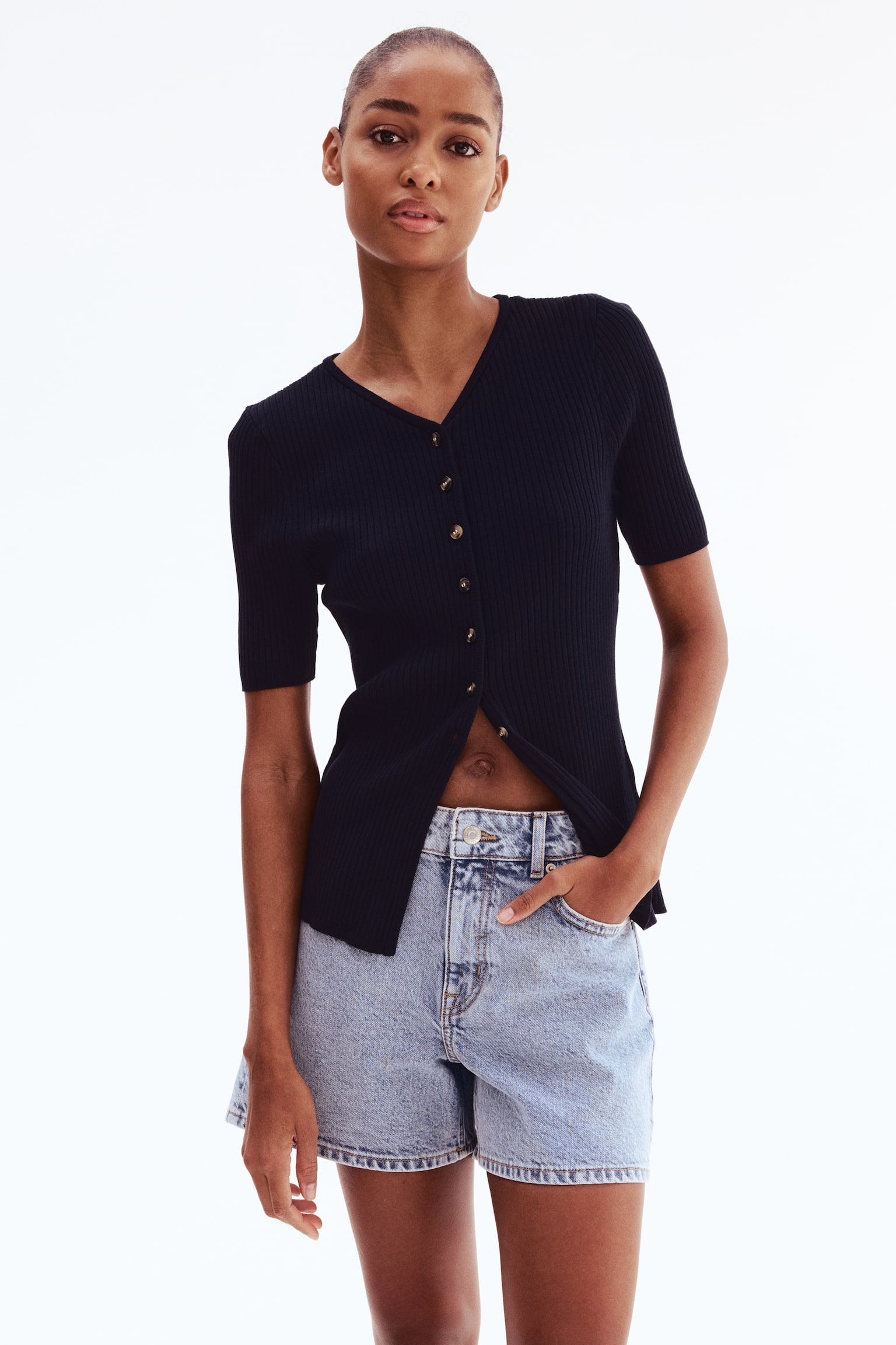 A long black rib-knit viscose-blend cardigan with a V-shaped neckline and short sleeves is being worn by the model. Buttons extend down to the waist, with several lower buttons left unfastened, revealing the model's midriff. The model is pairing the cardigan with light-wash denim shorts. - 1