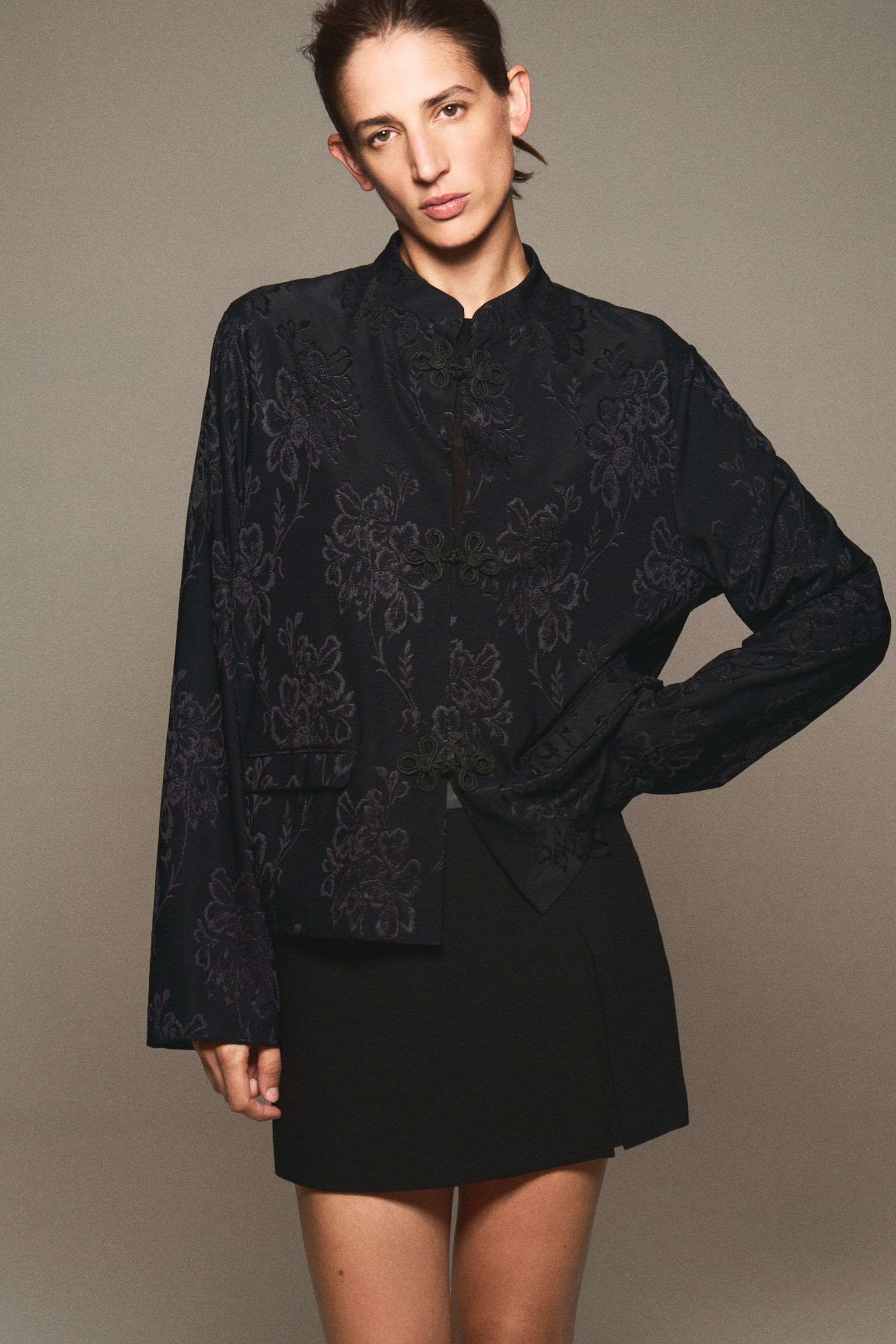 A black, loose-fit jacket in a tonal floral jacquard pattern is hanging facing forward, showcasing a band collar, dropped shoulders, and three decorative knot buttons down the centre. The jacket features long sleeves and jetted front pockets with flaps. - 1
