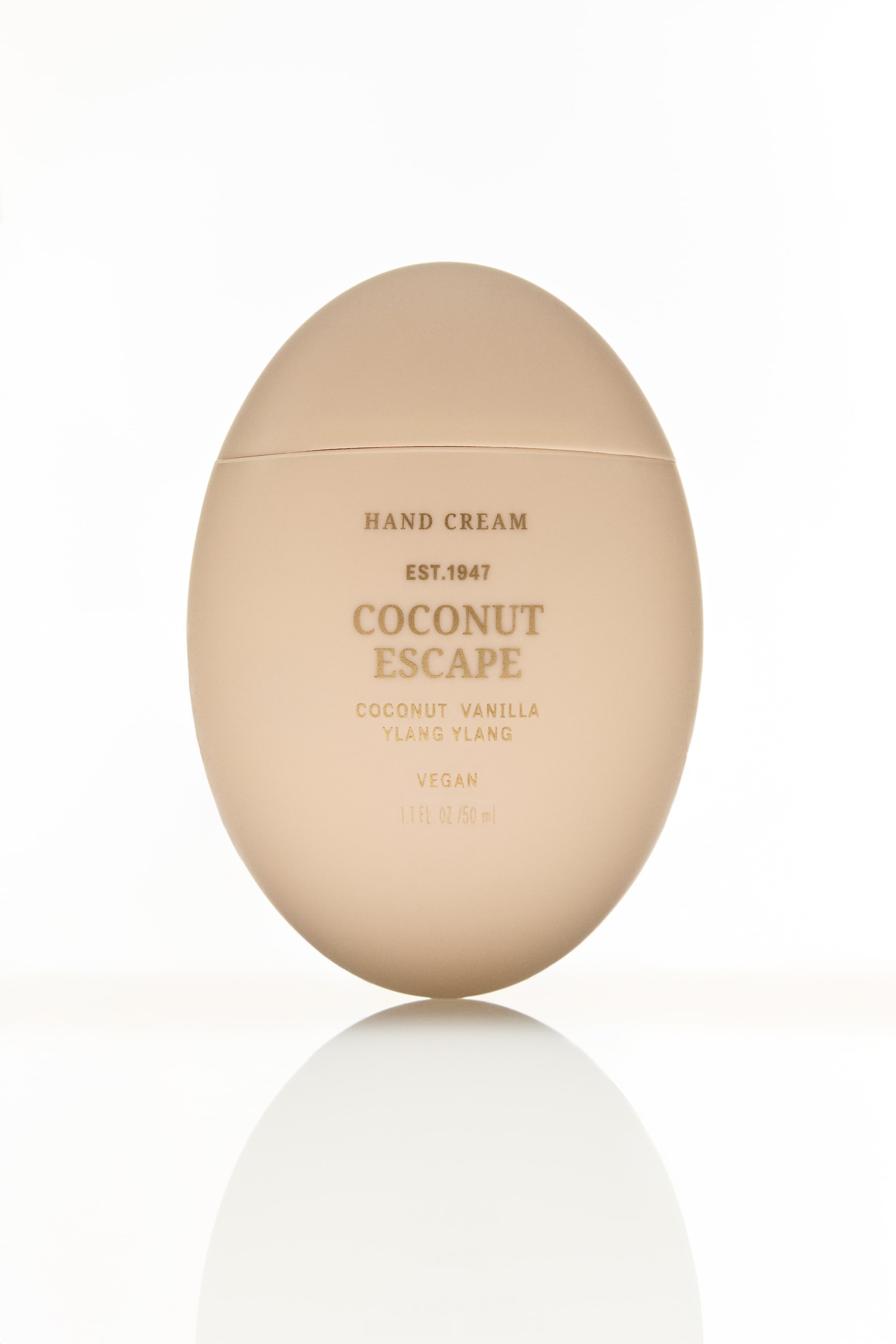 Ampliar la imagen: Un envase de crema de manos beige mate y ovalado está de pie y orientado hacia adelante. Letras doradas detallan su frente con "Coconut Escape", "Crema de Manos" y otra información del producto, y se puede ver una costura horizontal cerca de la parte superior.